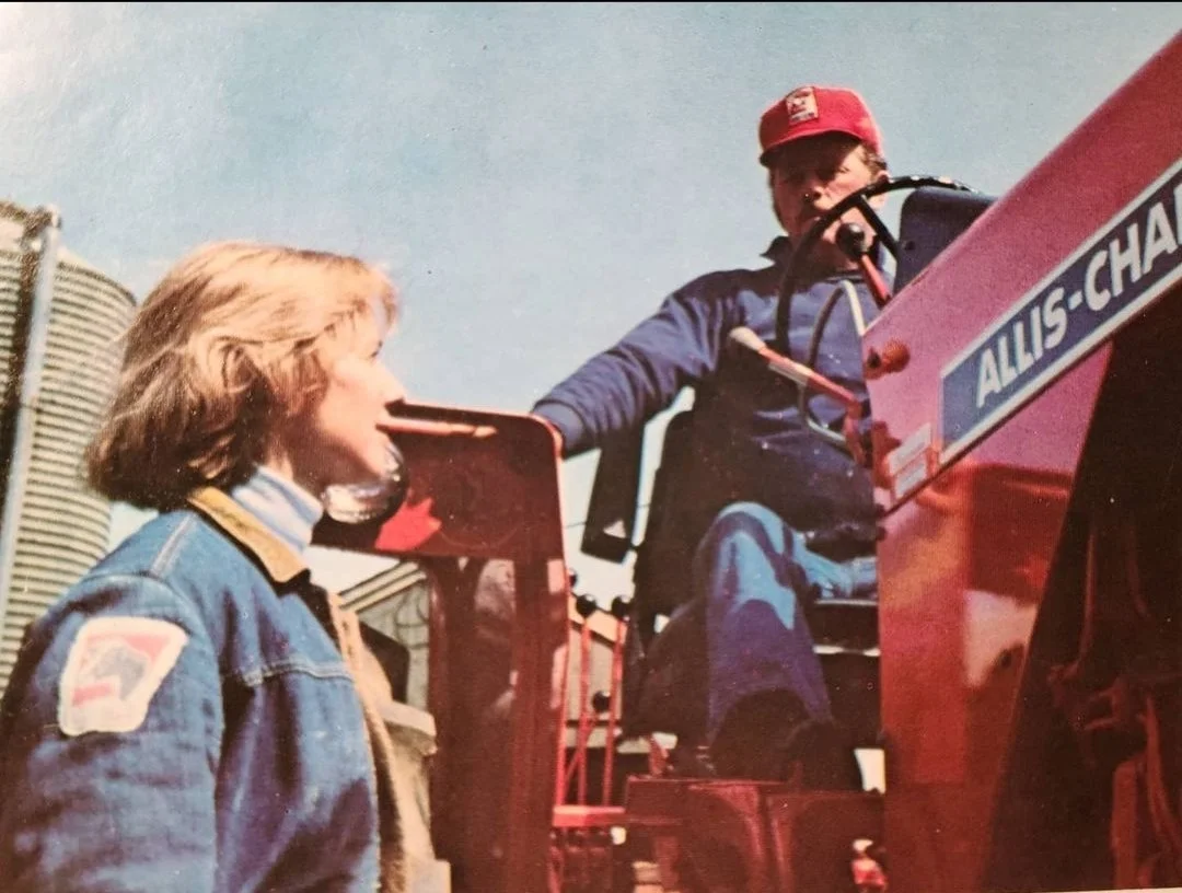 A young girl in a blue denim jacket talking to a boy sitting on a red Allis-Chalmers tractor, with a clear sky in the background.