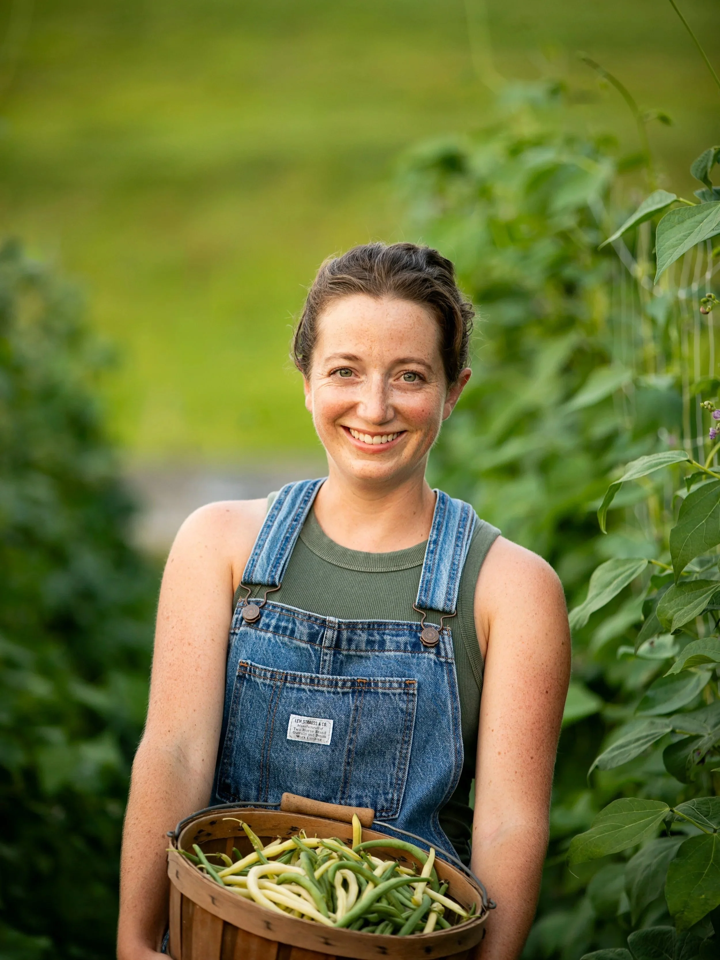 Woman smiling in a garden or farm holding a basket of freshly harvested green beans, surrounded by green plants.