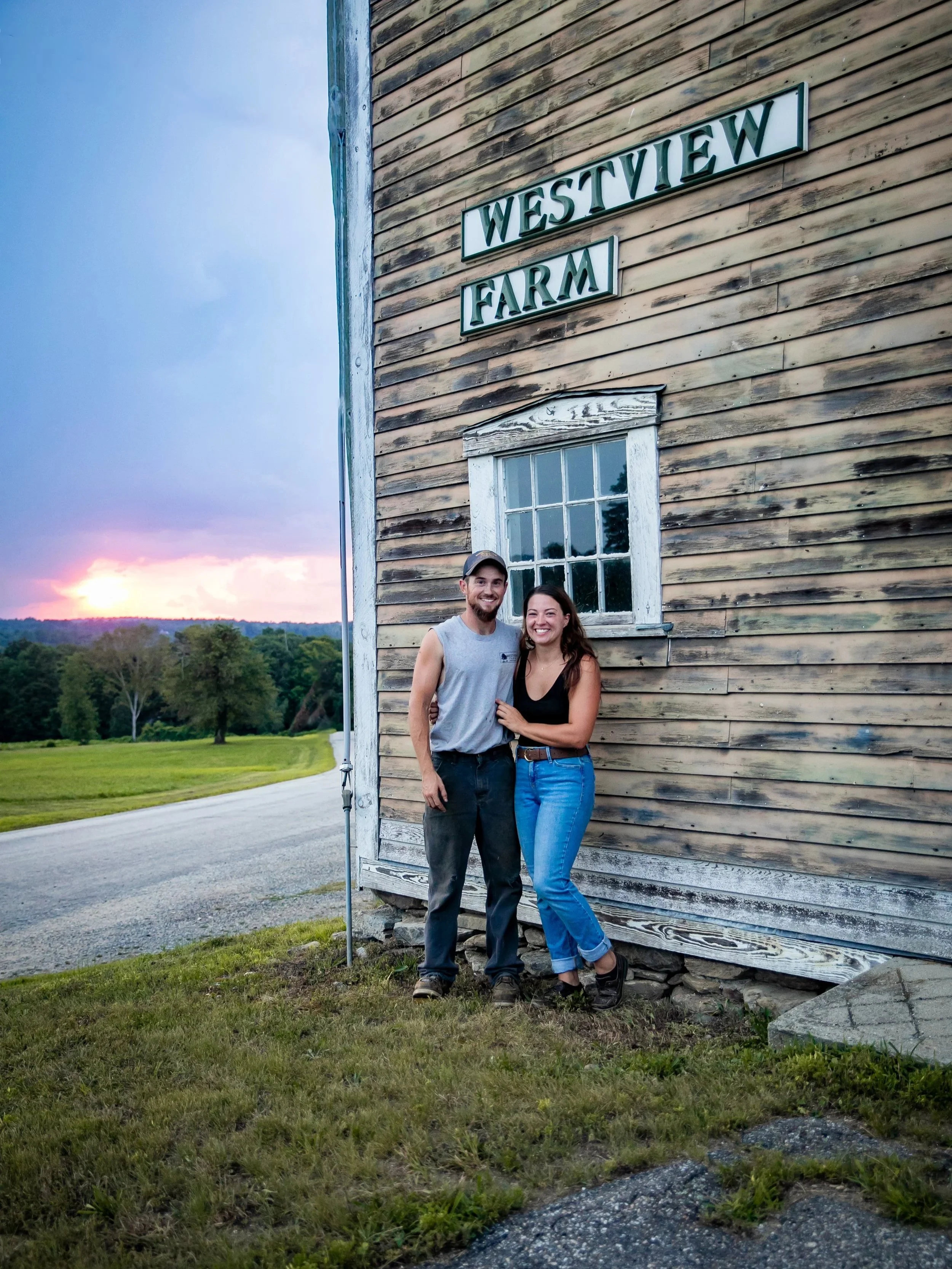 A couple standing outside a rustic wooden barn with a sign that reads 'Westview Farm' during sunset.