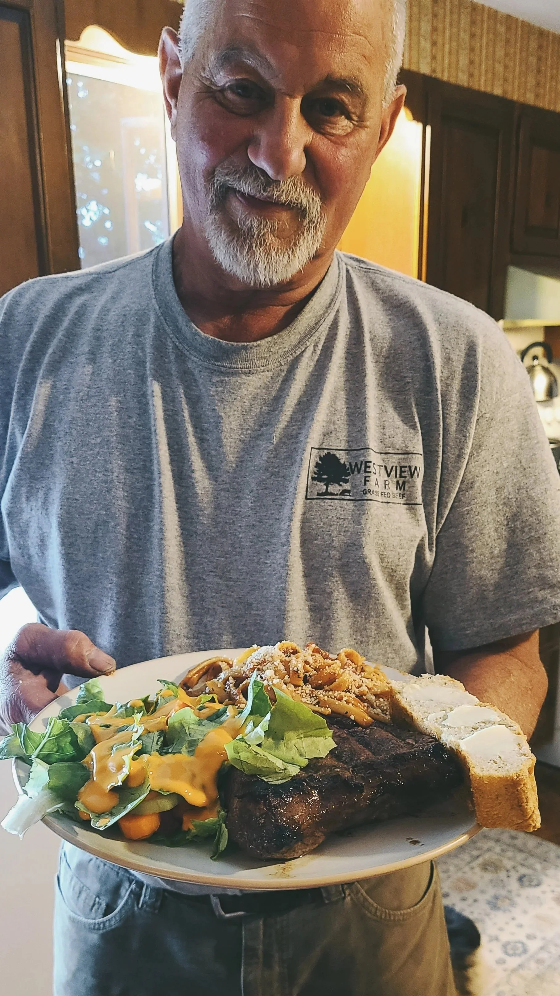 A man with white hair and beard, wearing a gray T-shirt, holding a plate with a meal that includes salad, a piece of grilled grass-finished beef, and cornbread, in a cozy kitchen setting.