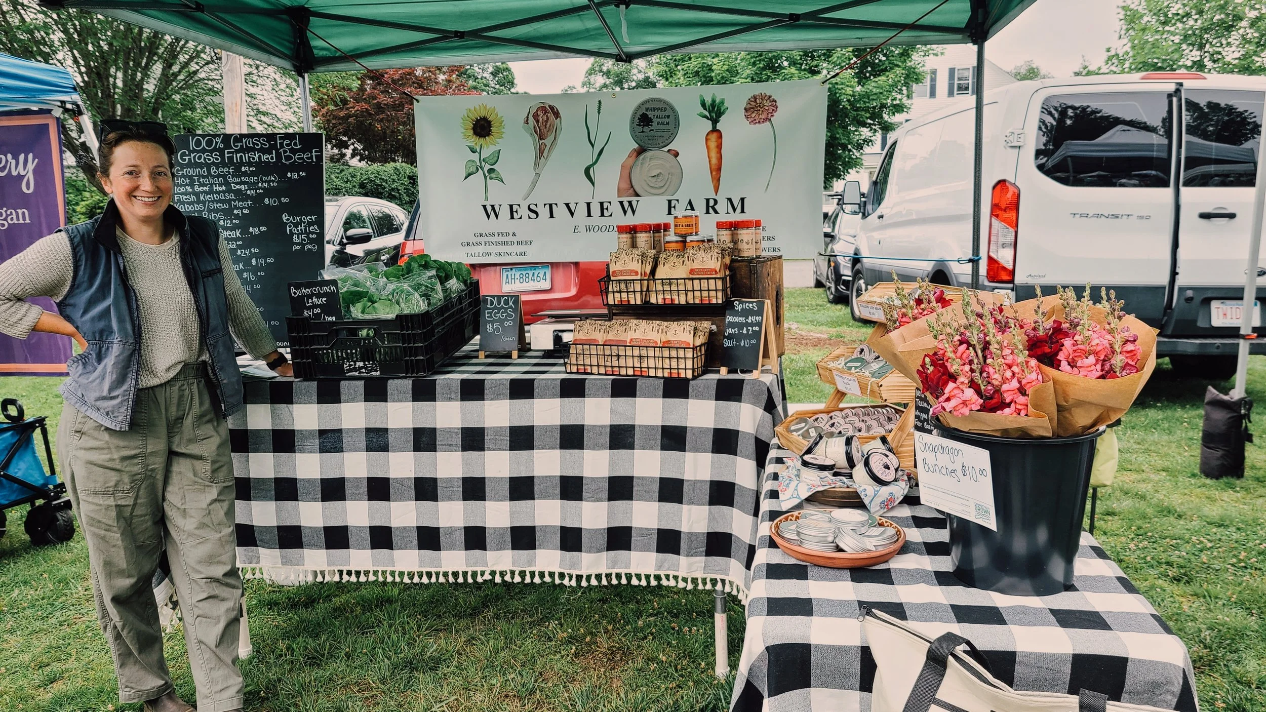 Woman smiling at a farmers market stall with fresh vegetables, herbs, and flowers, under a green canopy, with cars and trees in the background.
