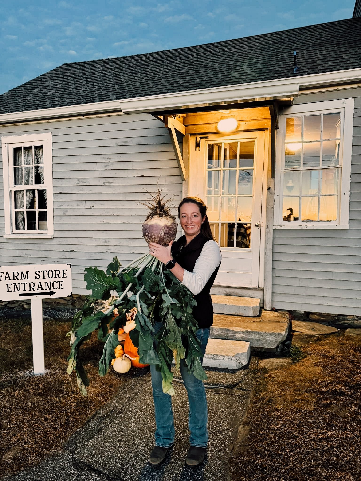 Woman standing outside a farm store entrance holding a massive root vegetable as big as her head, in front of a rustic building with a lit porch.