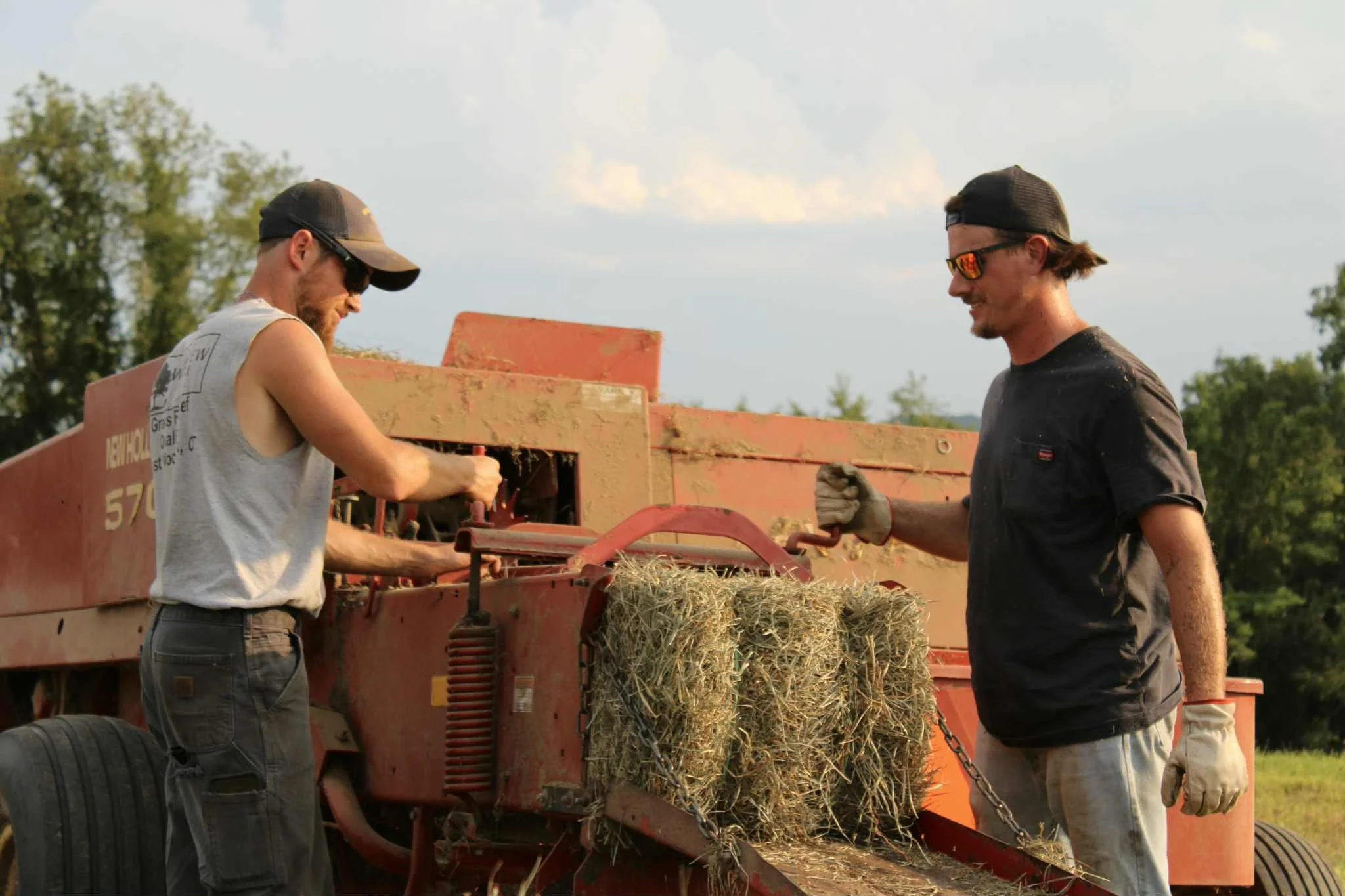 Two young men working on at Westview Farm with a hay baler one wearing a backwards cap and sunglasses, the other in gloves, with hay bales in the foreground and trees in the background.