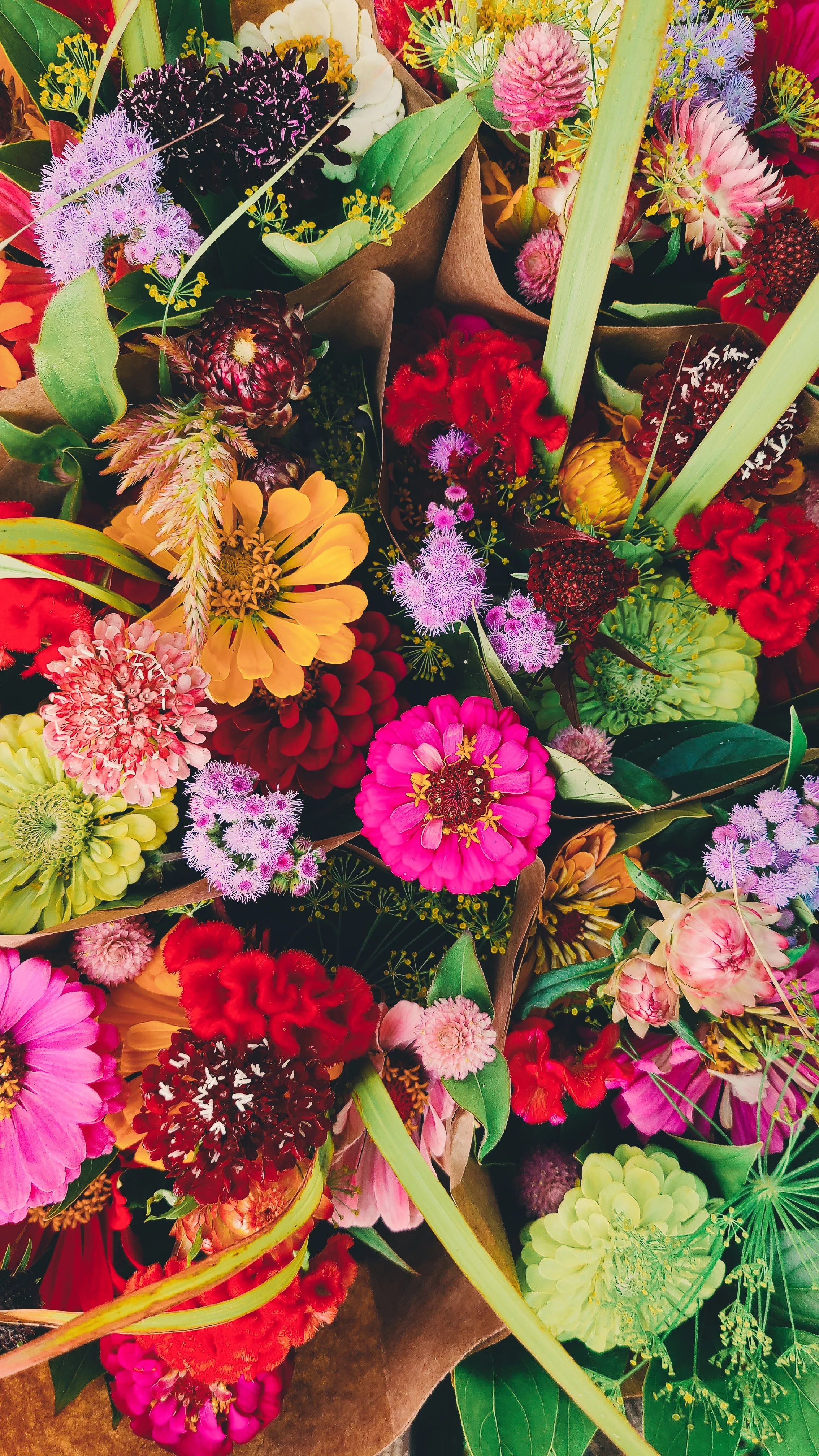 A vibrant bouquet of various colorful flowers including pink, red, yellow, purple, and green blossoms with green leaves and stems, wrapped in brown paper.