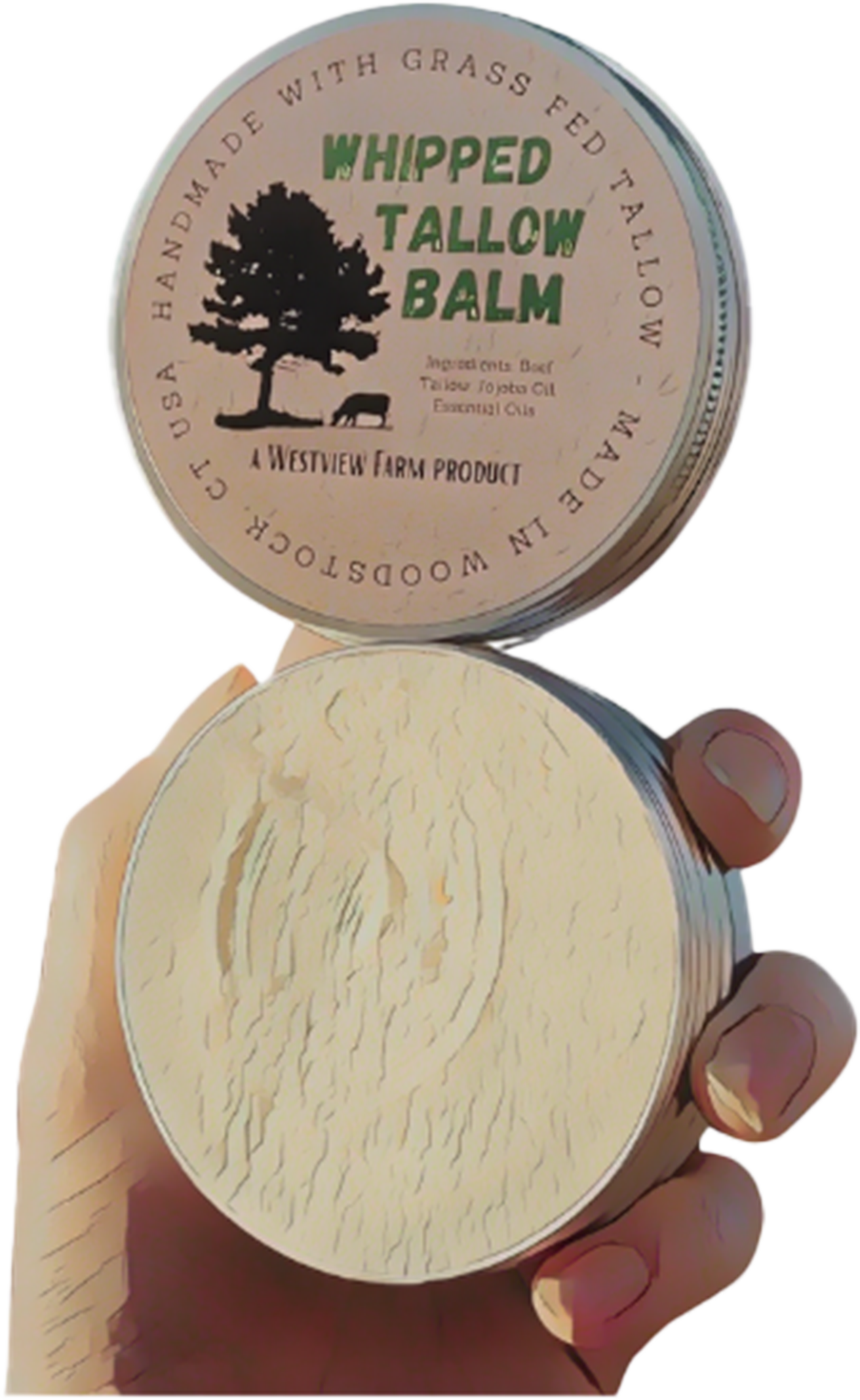 A hand holding a round tin of Whipped Tallow Balm from Westview Farm, with the lid off revealing the balm inside. The lid has a label with a tree silhouette, a pig silhouette, and text indicating the balm is made with grass-fed tallow, and listing ingredients such as beef, tallow, jojoba oil, and essential oils.