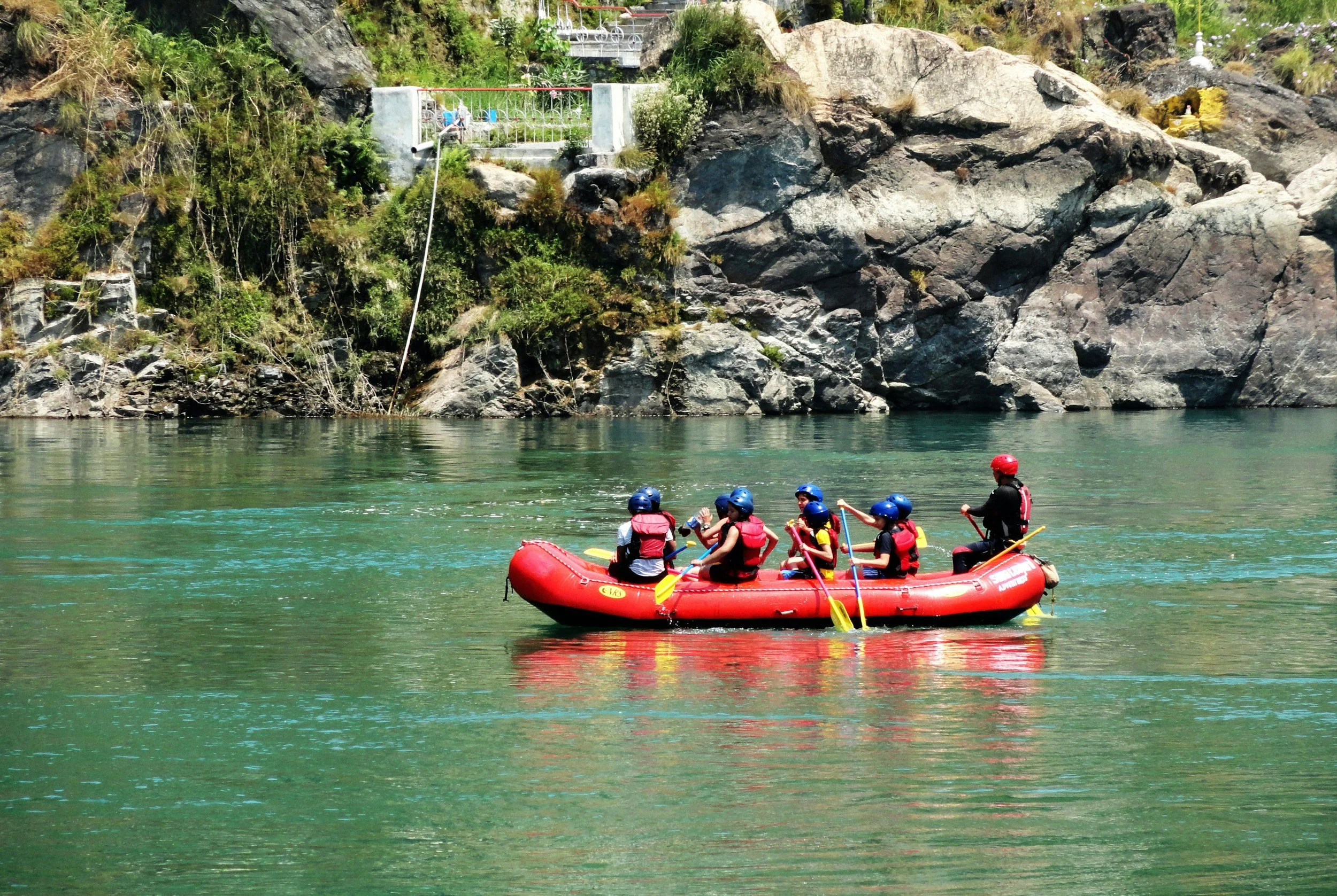 Groupe de personnes en rafting sur une rivière verte, portant des casques bleus et rouges, avec des gilets de sauvetage, près d'une falaise rocheuse avec de la végétation.