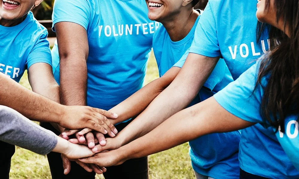 Groupe de personnes portant des T-shirts bleus avec l'inscription 'BÉNÉVOLE', qui se tiennent par la main en cercle, probablement lors d'une activité de bénévolat en extérieur.