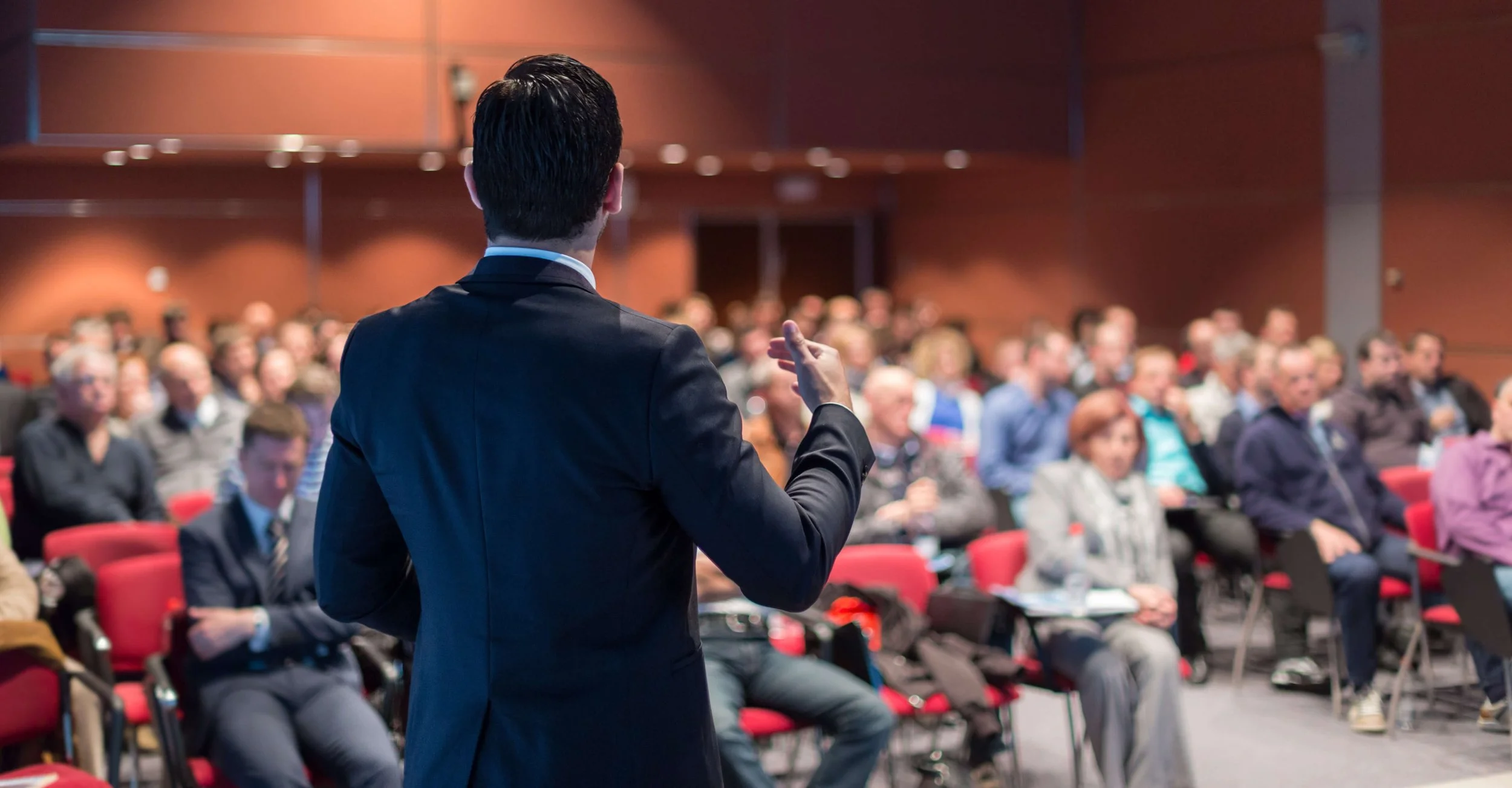 Un homme en costume noir parle à un auditoire dans une salle de conférence avec des sièges rouges. La salle est pleine de personnes assises et écoutant attentivement.