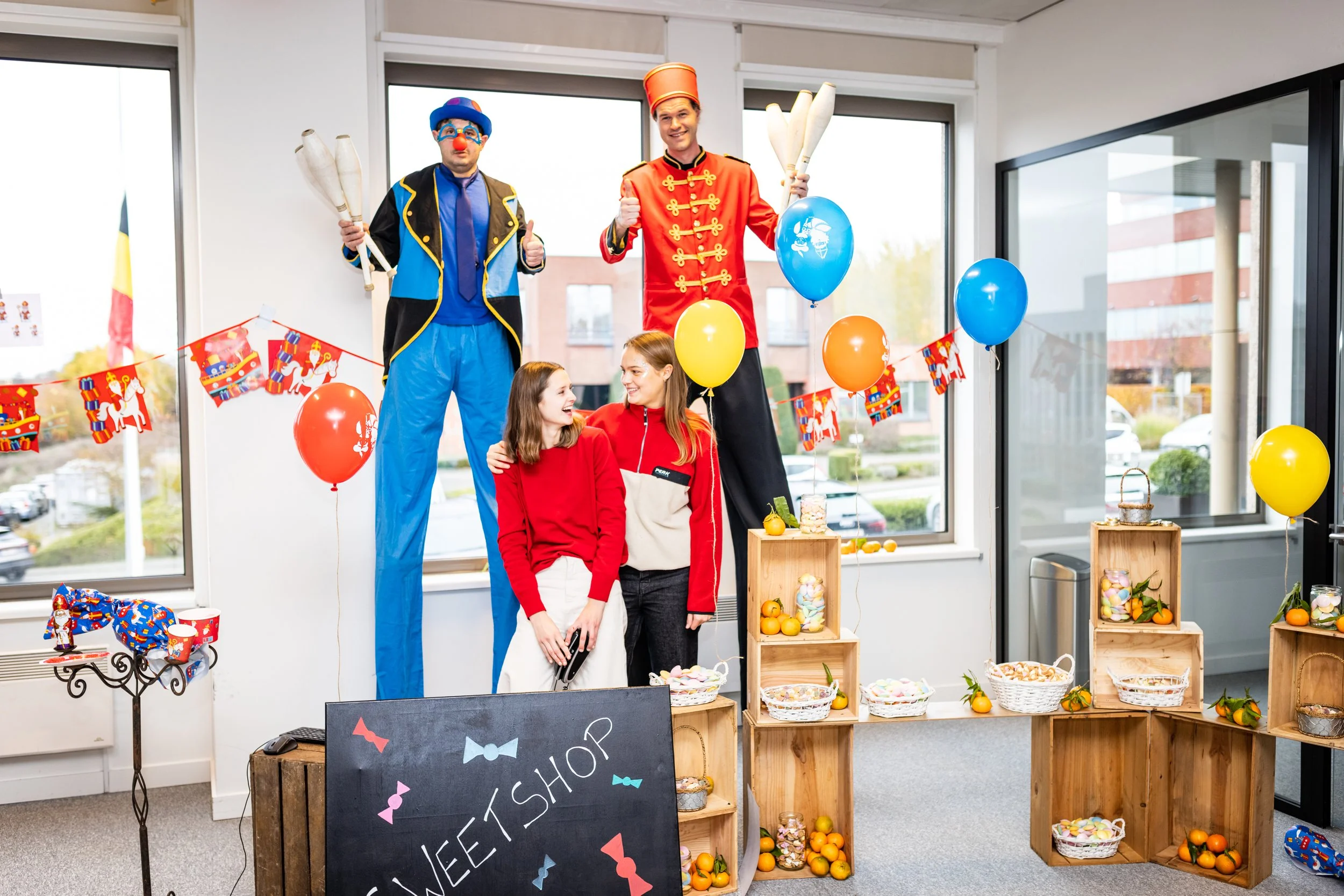 Une scène de fête avec deux hommes habillés en clowns et deux jeunes filles souriantes devant un stand décoré avec des ballons, des oranges, et des bonbons, dans une pièce lumineuse avec de grandes fenêtres.