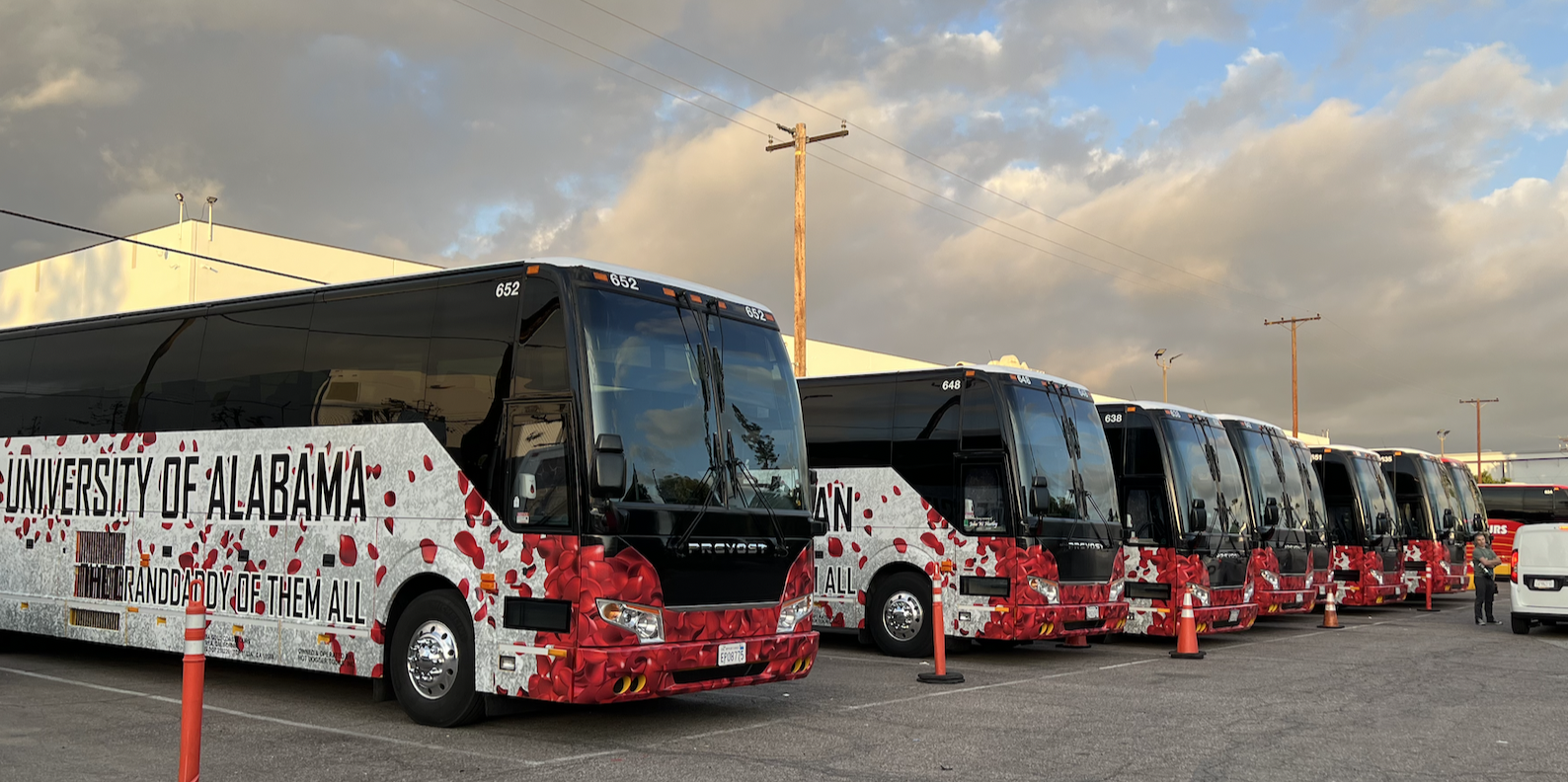 A line of University of Alabama buses decorated with crimson and white flowers parked in a lot under a cloudy sky.