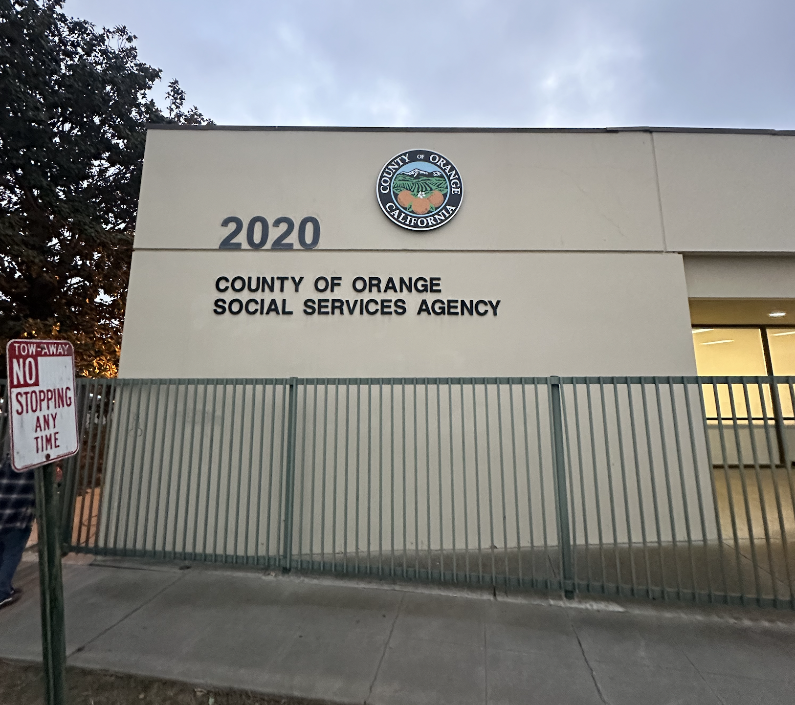 Exterior of the County of Orange Social Services Agency building in California with a sign indicating the address 2020, featuring the California and Orange County seal at the top.