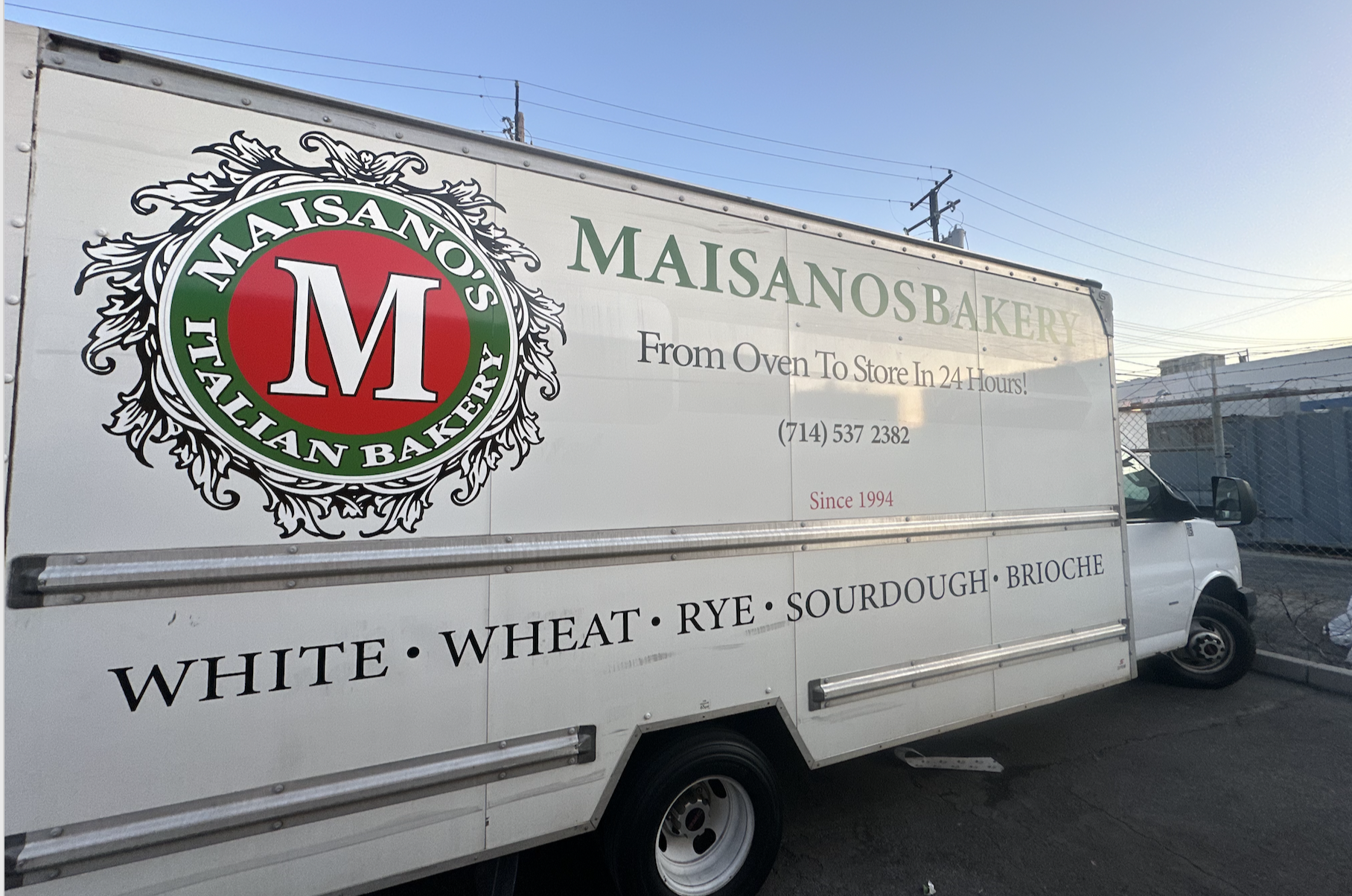 Delivery truck with Masanos Bakery logo and text advertising Italian baked goods, with a clear sky and utility poles in the background.