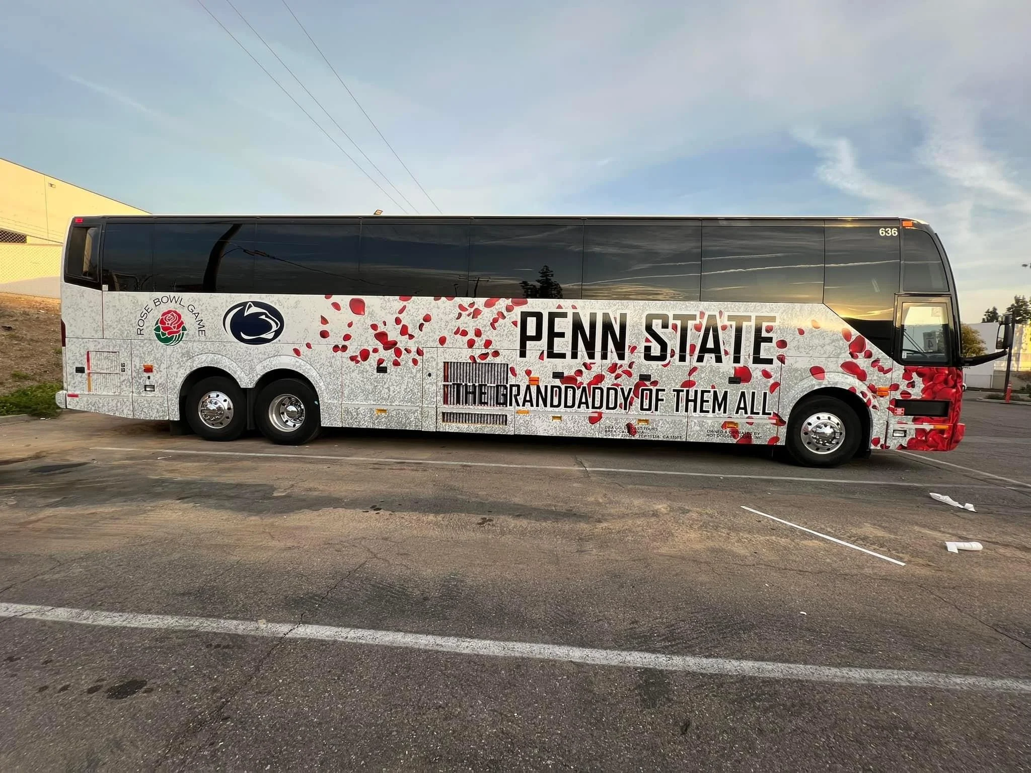 A Penn State tour bus with branding for Rose Bowl game and The Granddaddy of Them All. The bus has a white background with red rose petal graphics and logos, including the Penn State logo and Rose Bowl logo, parked on a paved lot during sunset.