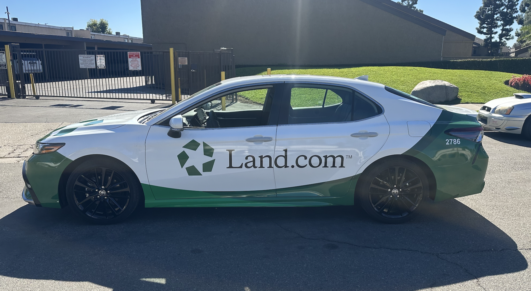 A white and green Land.com promotional car with black wheels parked in a lot, with a gated parking area and a grassy hill in the background.