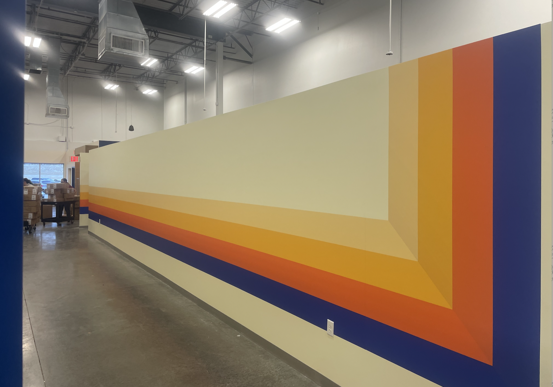 Interior of a warehouse with a colorful mural on the wall featuring a geometric, layered design in shades of yellow, orange, red, and blue.