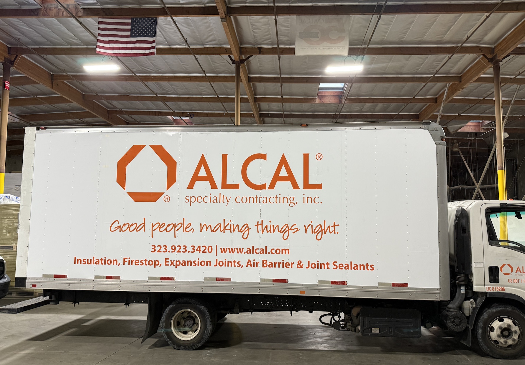 A delivery truck inside a warehouse with a white body and orange lettering advertising ALCAL specialty contracting, Inc., with their logo, contact information, website, and a list of services including insulation, firestop, expansion joints, air barrier, and joint sealants. An American flag hangs from the ceiling above.