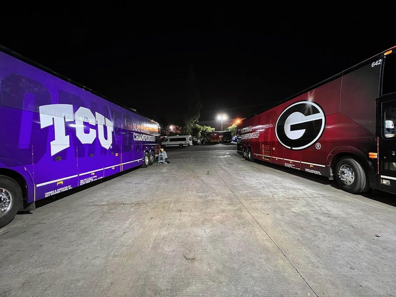 Nighttime parking lot with two large buses, one purple with 'TCU' and 'Championship' written on it, the other red with a large black 'G' logo, and a few vehicles in the background.