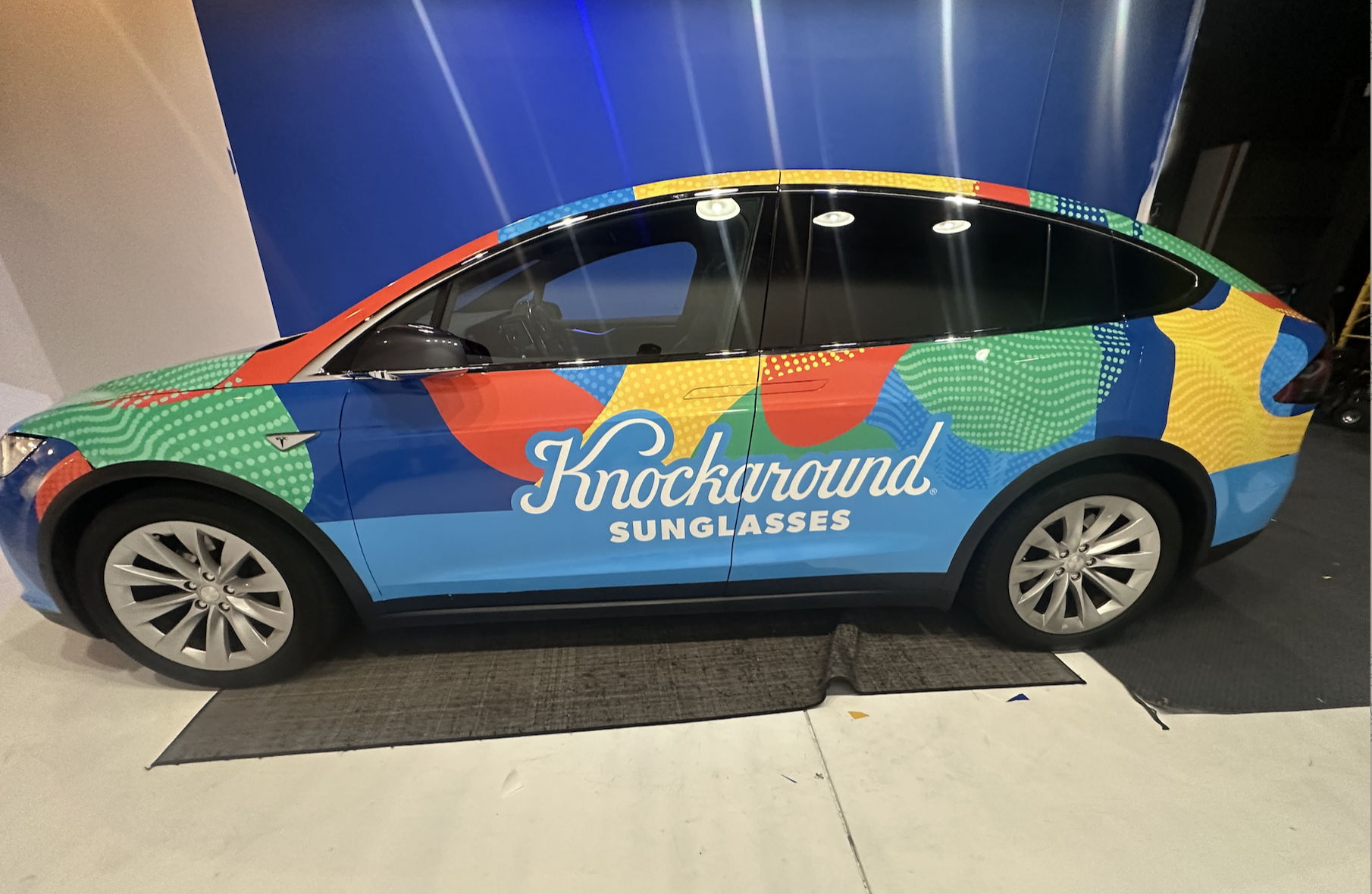 Shaded electric car with colorful advertisements reading 'Knockaround Sunglasses' parked indoors, with a blue backdrop and ceiling lights overhead.