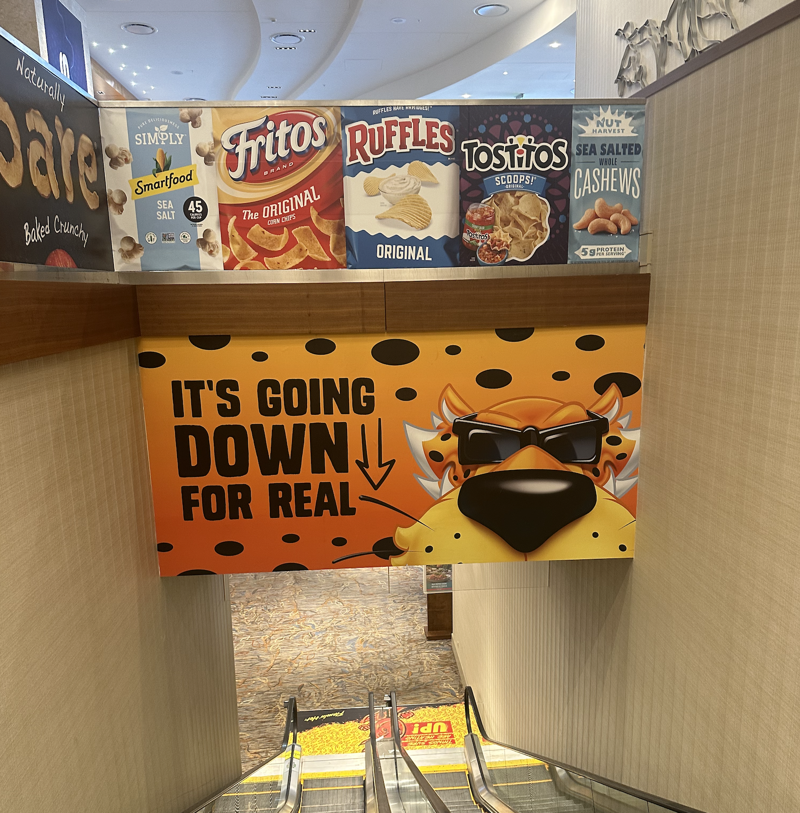 Colorful aisle sign with tiger cartoon wearing sunglasses and a yellow bandana, saying "It's going down for real" with black dots on an orange background, located near escalators in a shopping mall.