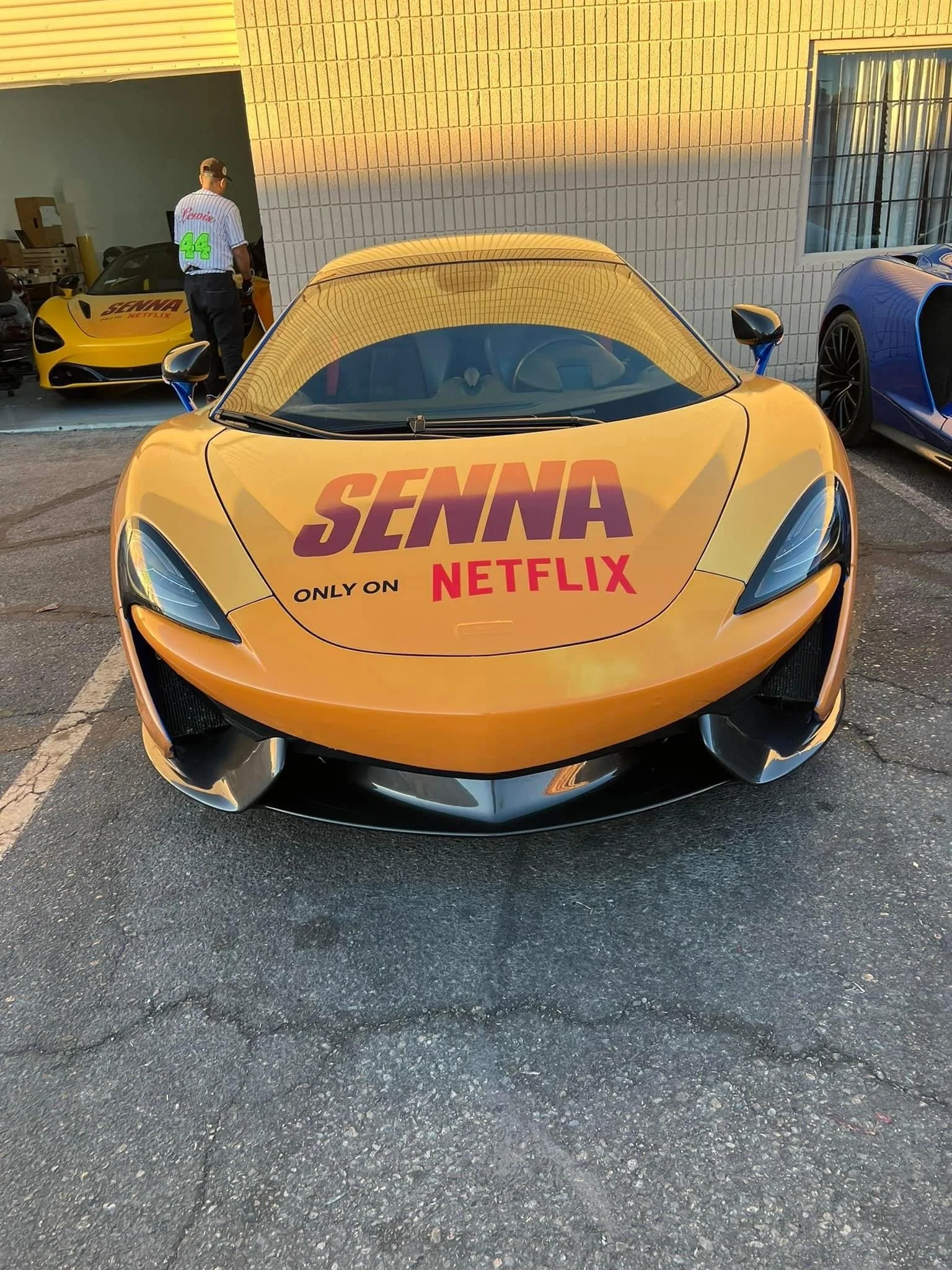 A yellow McLaren sports car with a promotional wrap featuring the word 'SENNA' in large purple letters, 'ONLY ON NETFLIX' below in smaller black and red text, parked in a parking lot. Other sports cars and a person standing near a yellow race car are visible in the background.
