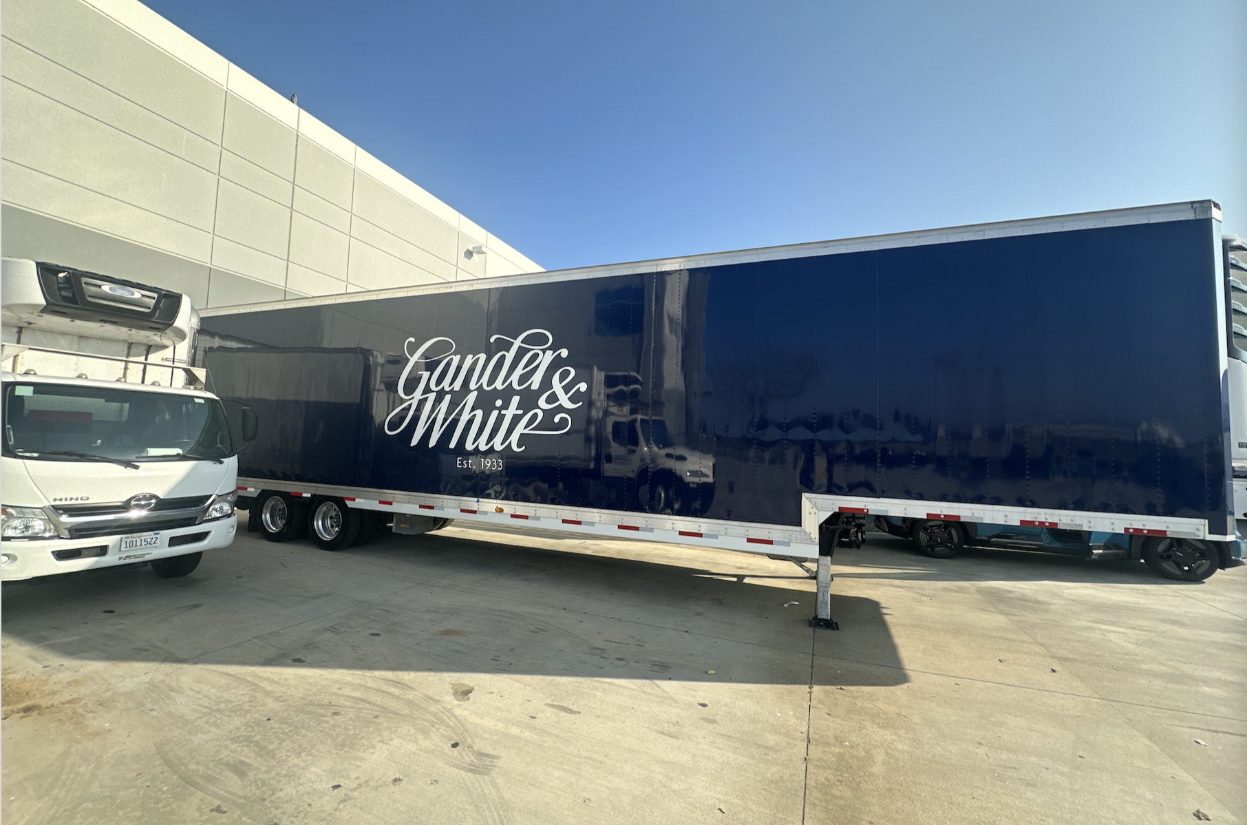 A large Gander & White shipping trailer in navy blue with white cursive lettering, parked on a concrete lot beside a white truck, against a clear blue sky and a beige building.