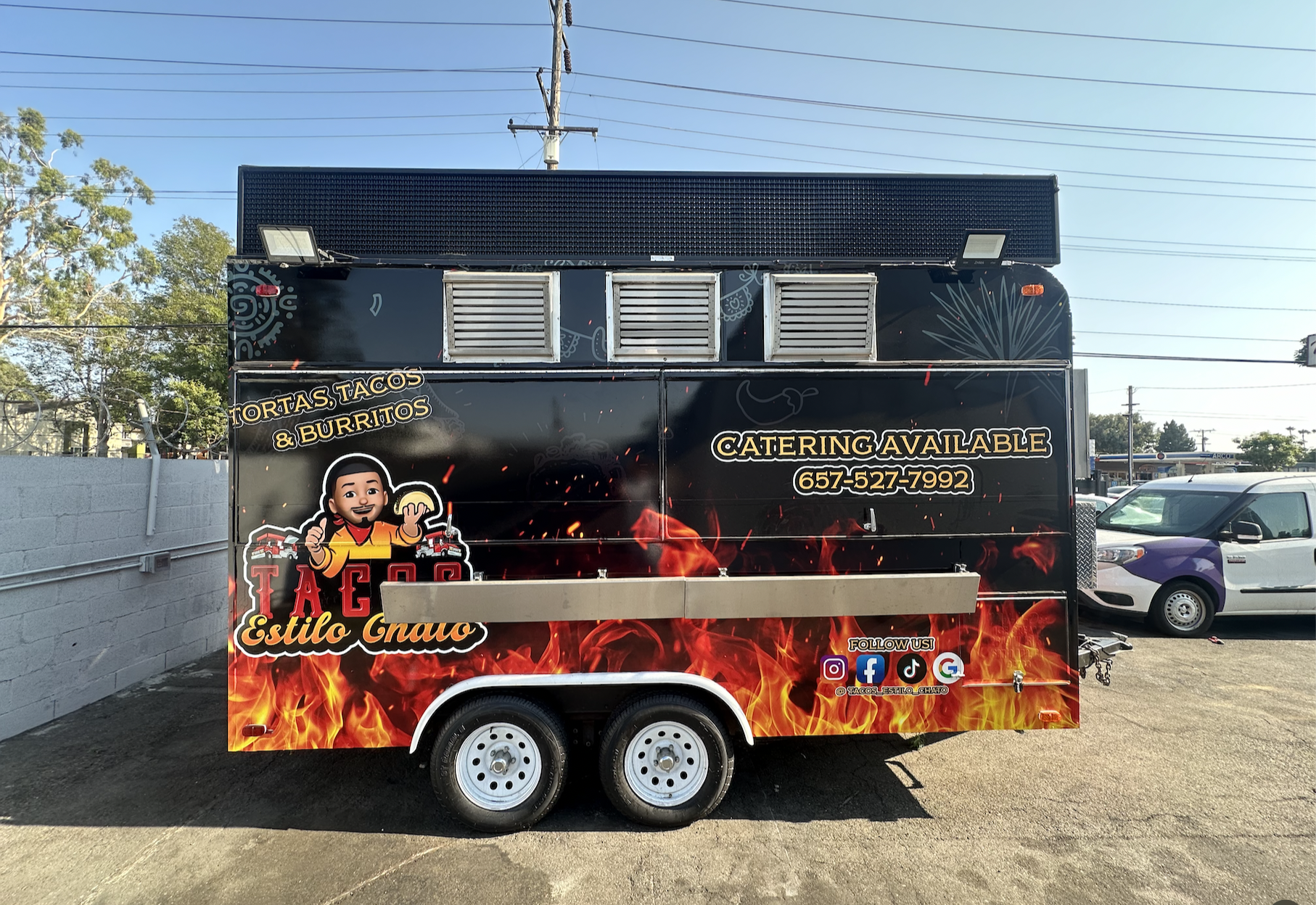 A food truck with black and red flames graphics, advertising tacos and burritos with a phone number and social media icons.