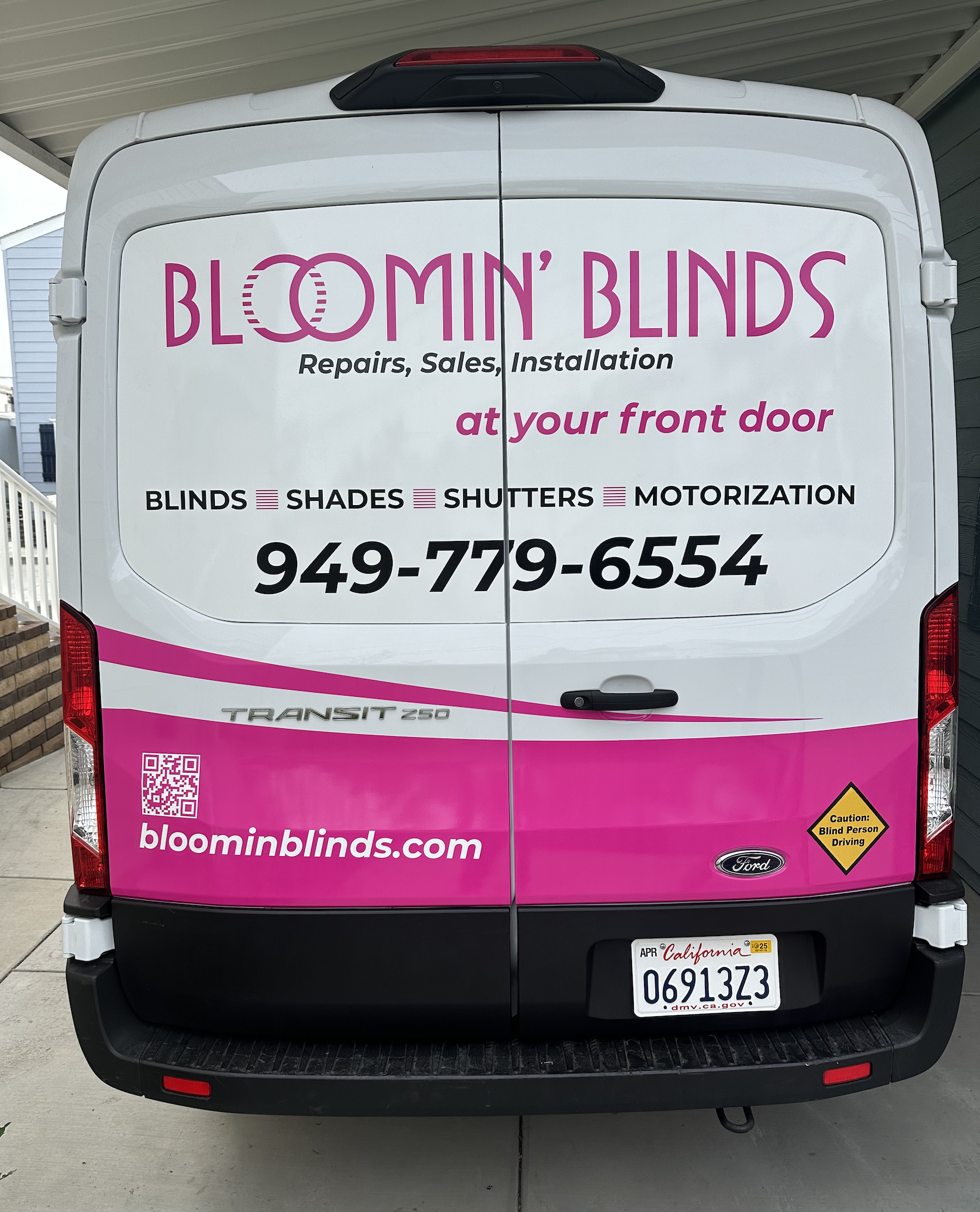 The back of a white Ford Transit 250 van with pink and black lettering advertising Bloomin' Blinds, a company offering repairs, sales, and installation of blinds, shades, shutters, and motorization, with a phone number, website, and a caution sign for blind person driving.