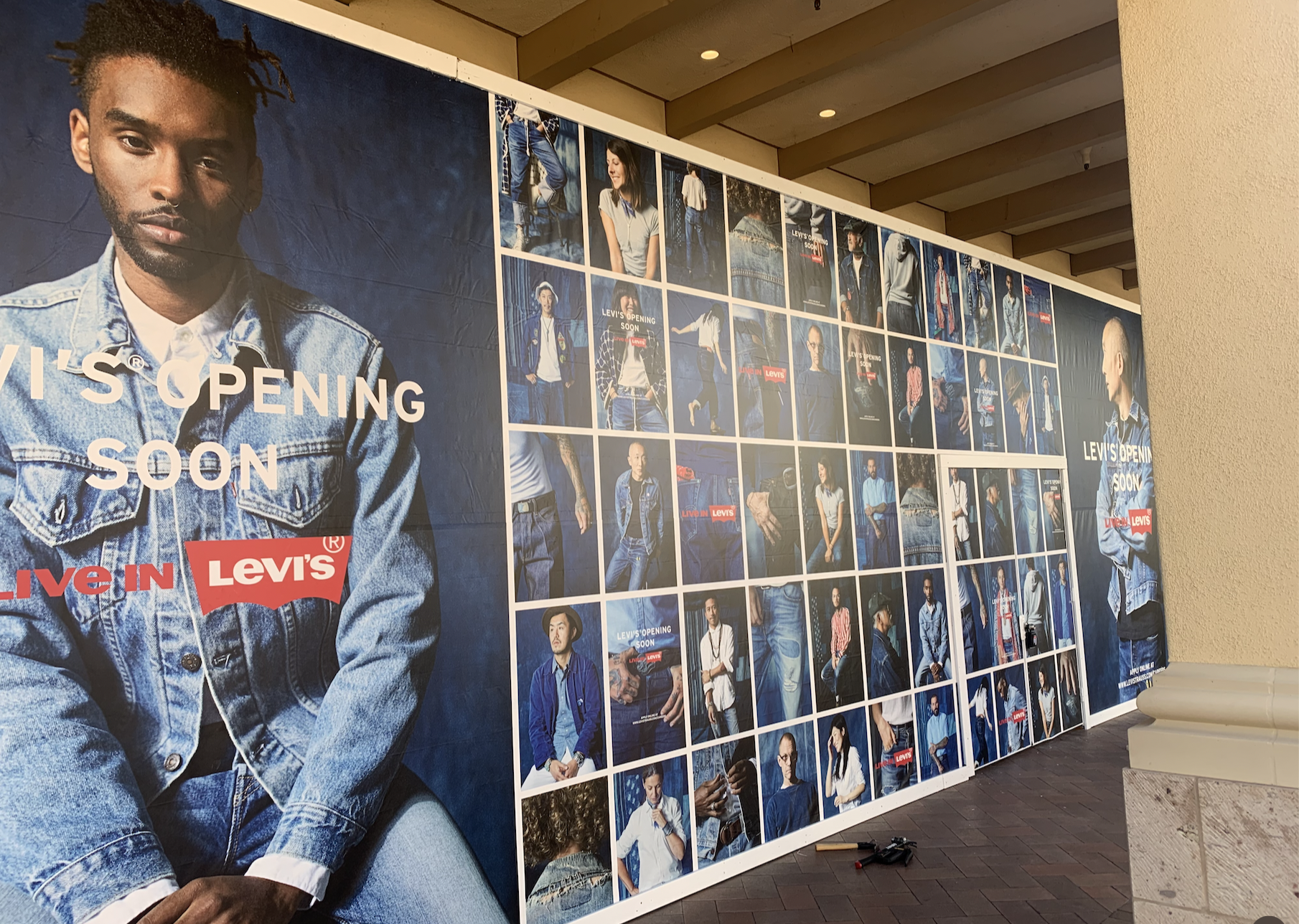 Large promotional board for Levi's clothing store opening soon, featuring a variety of model photos showcasing denim outfits, with larger images of male and female models on either end, and smaller photos in the center.