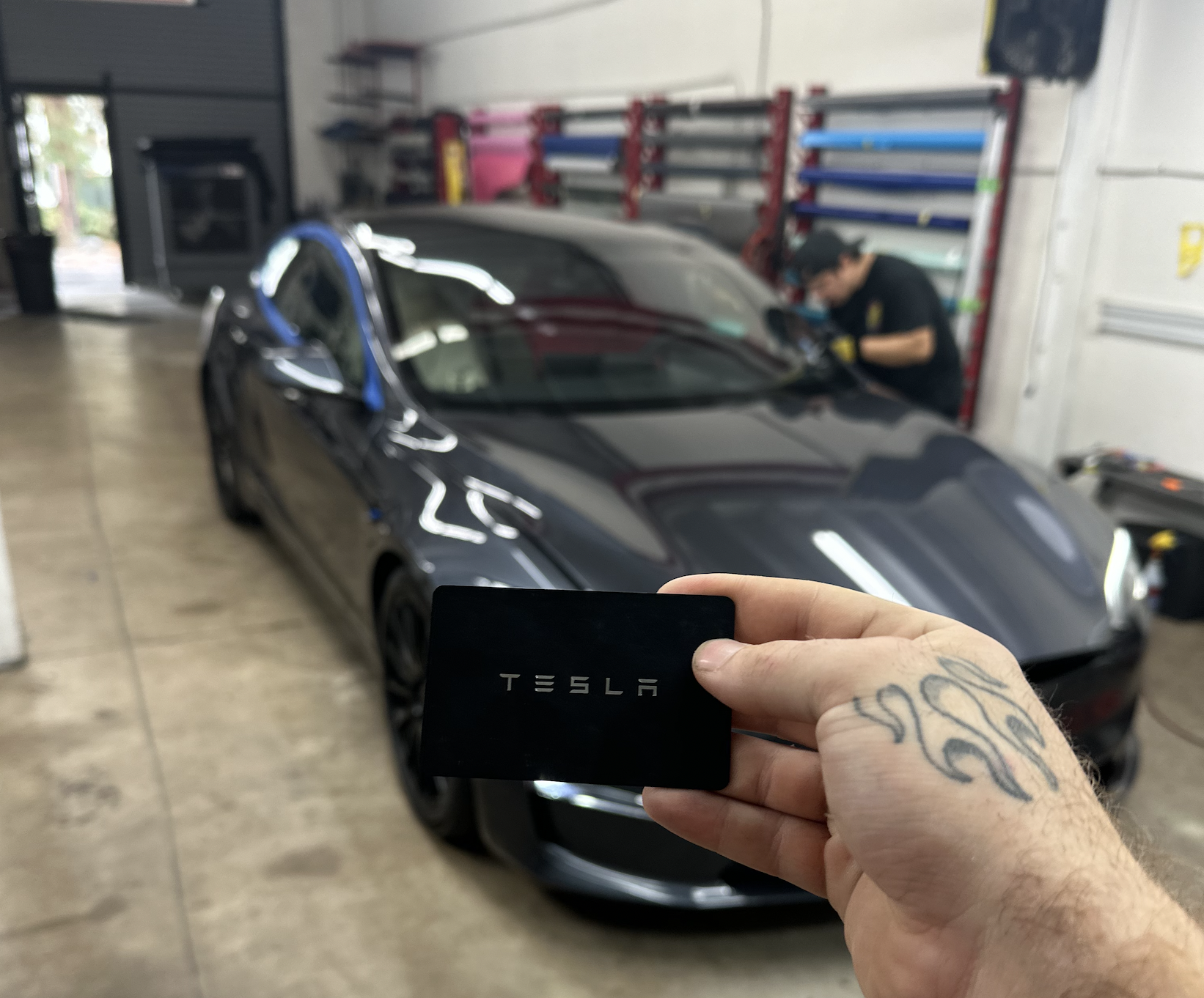 Person holding a Tesla card in front of a black Tesla sports car inside a garage, with a mechanic working on the car in the background.