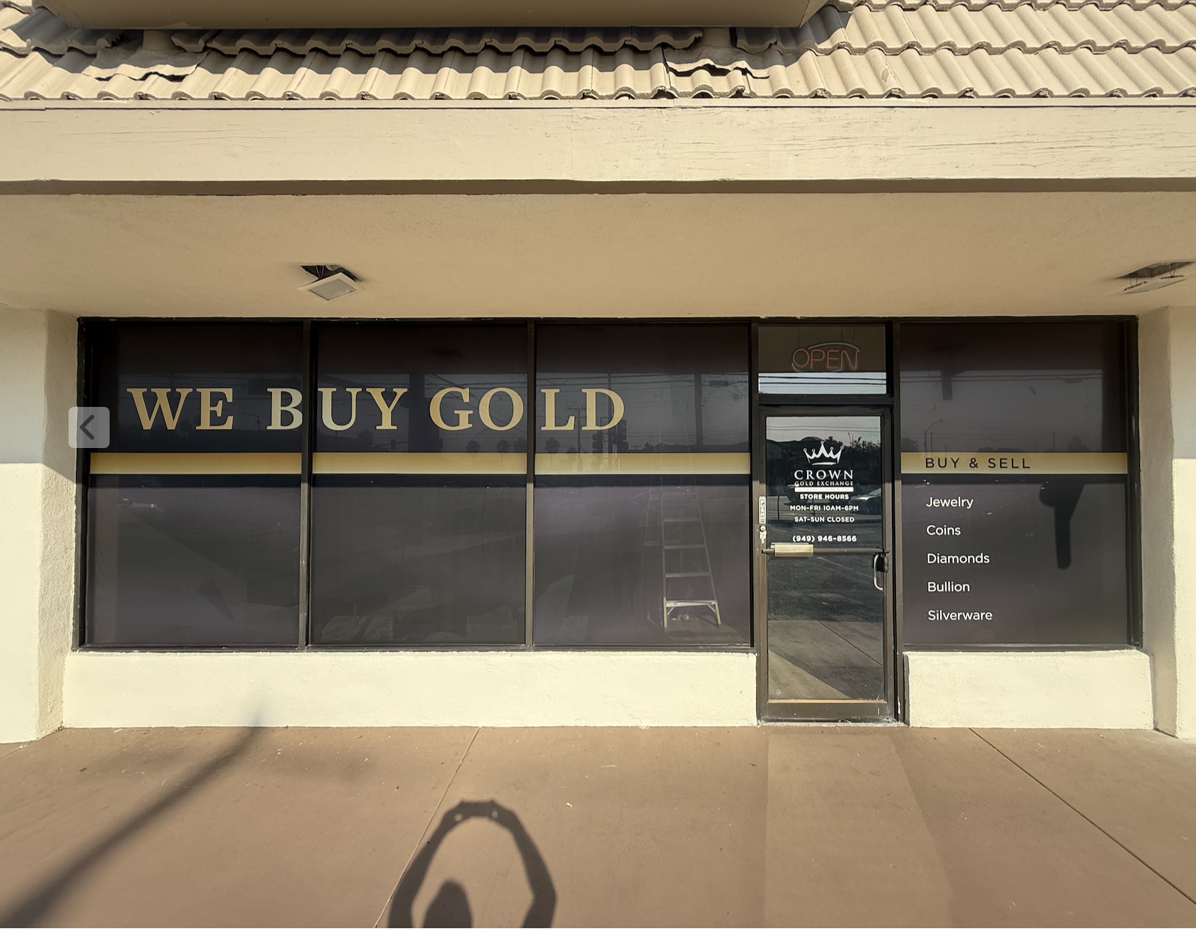 Storefront with large windows displaying the words 'WE BUY GOLD' and 'BUY & SELL'. The door has an 'OPEN' sign and lists store hours. The store buys and sells jewelry, coins, diamonds, bullion, and silverware.