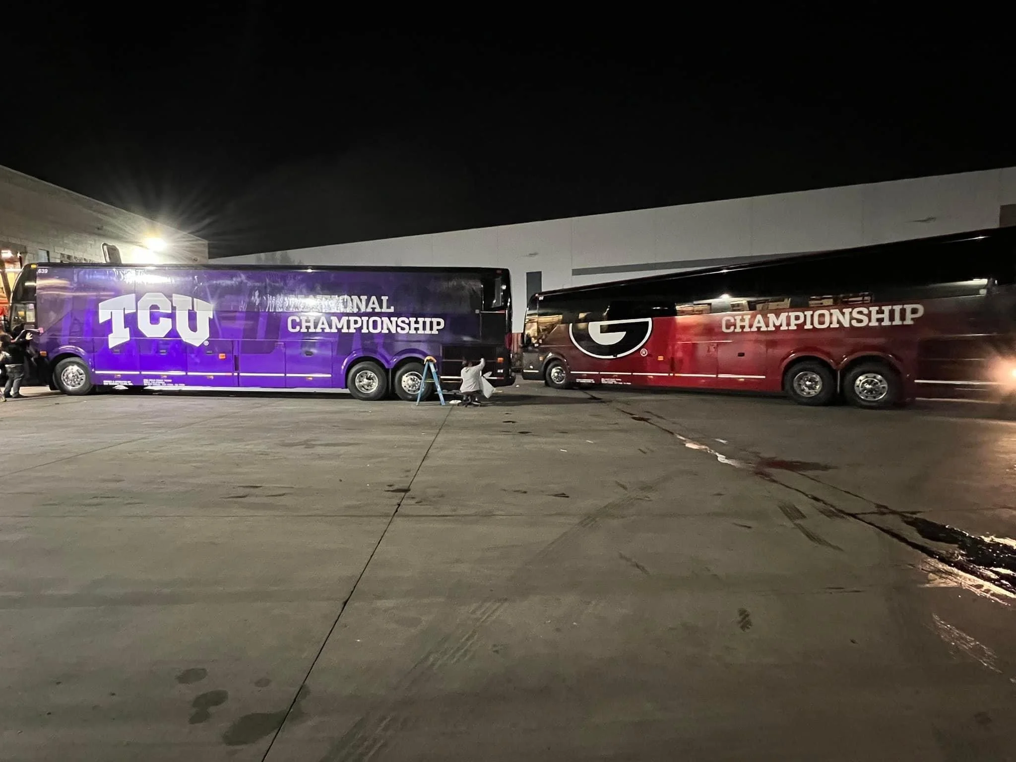 Two bus trailers at night, one purple with 'TGU NATIONAL CHAMPIONSHIP' and one red with a large G and 'CHAMPIONSHIP', parked outside a building with bright overhead lights.