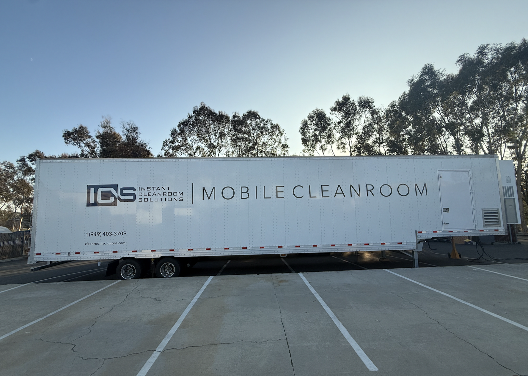 A large white mobile cleaning unit with the logo and contact information for IQS Instant Cleanroom Solutions, parked in a parking lot with trees in the background.
