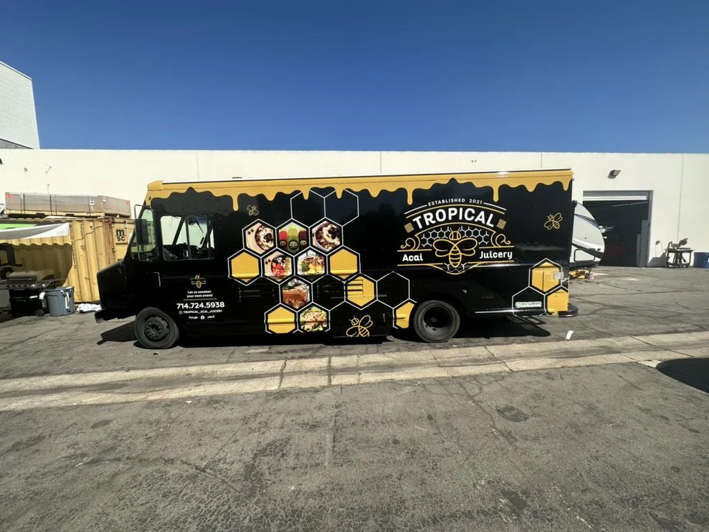 A black food truck with yellow and white accents parked outside a building, advertising Tropical Acai Juicery with images of acai bowls and honeycomb patterns.