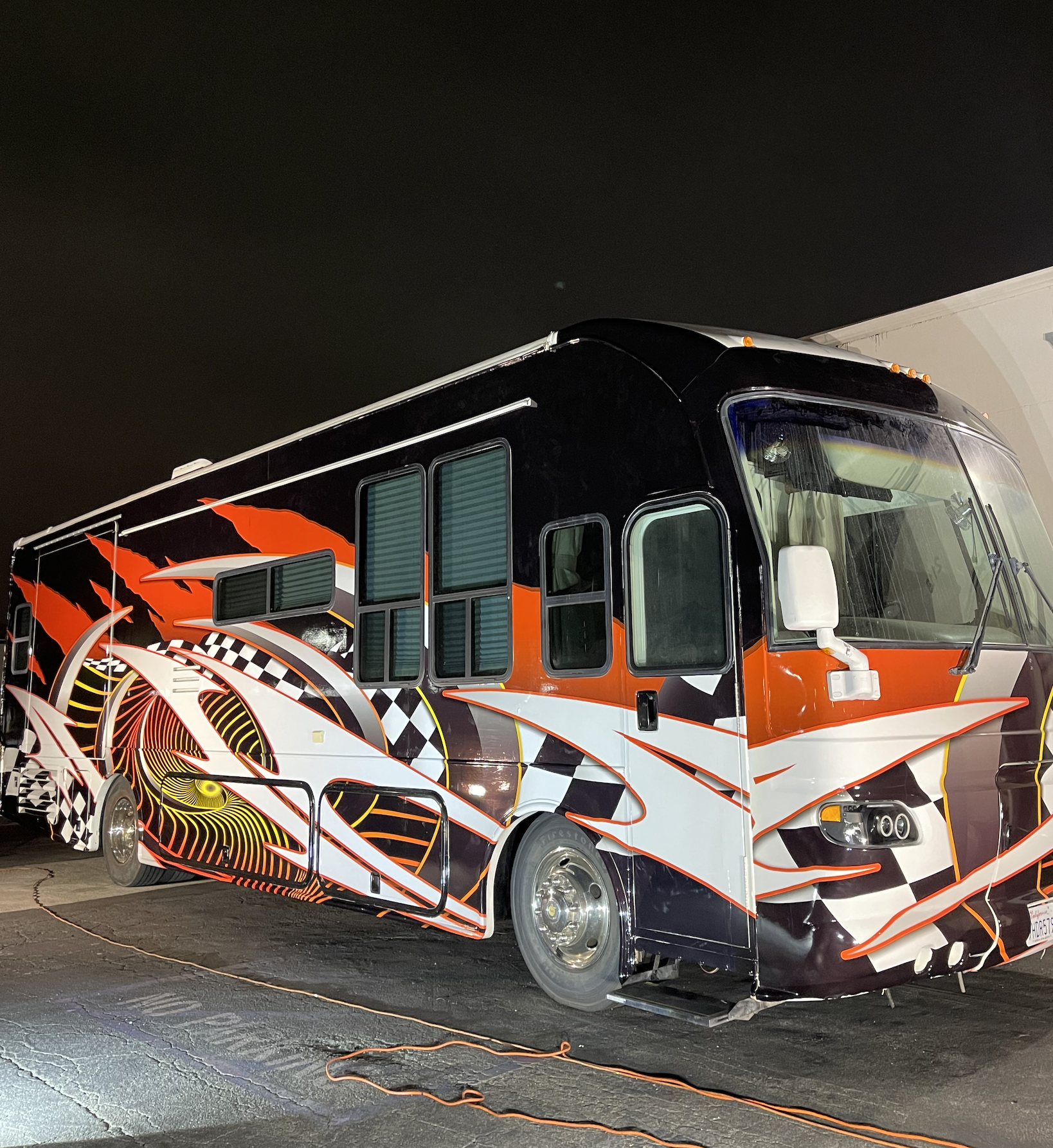 A painted racing-themed bus parked at night with black, white, orange, and yellow flame and checkered patterns on the body.