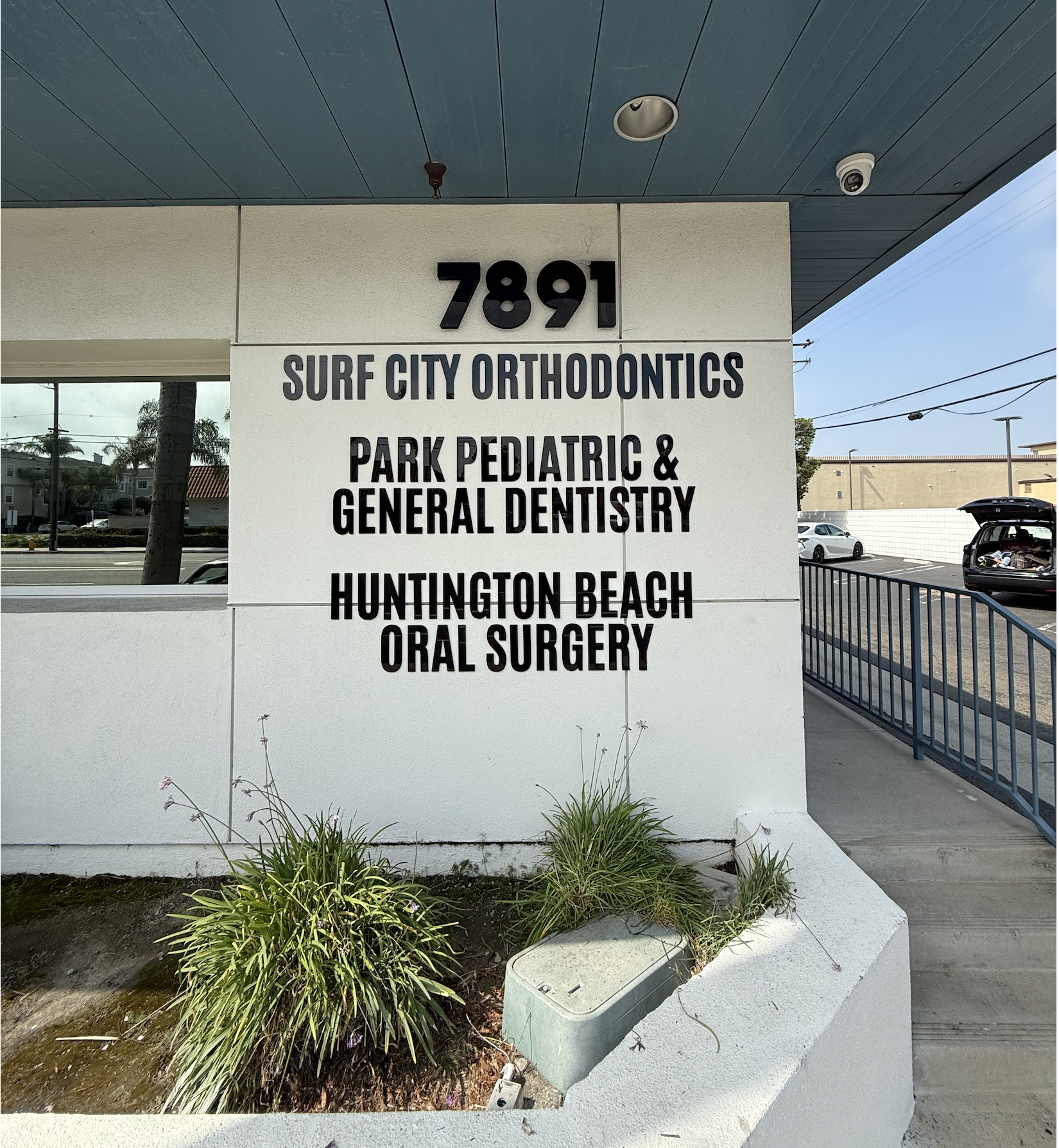 Sign with the address 7891 listing Surf City Orthodontics, Park Pediatric & General Dentistry, Huntington Beach Oral Surgery, on a white wall with plants and a parking lot in the background.