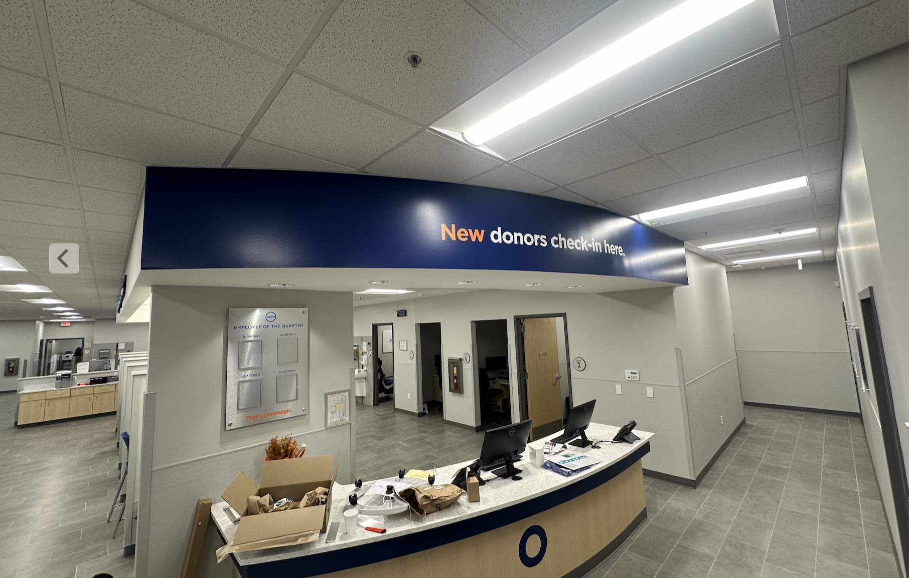 Hospital reception area with a sign indicating 'New donors check-in here' and a counter with computers and office supplies.