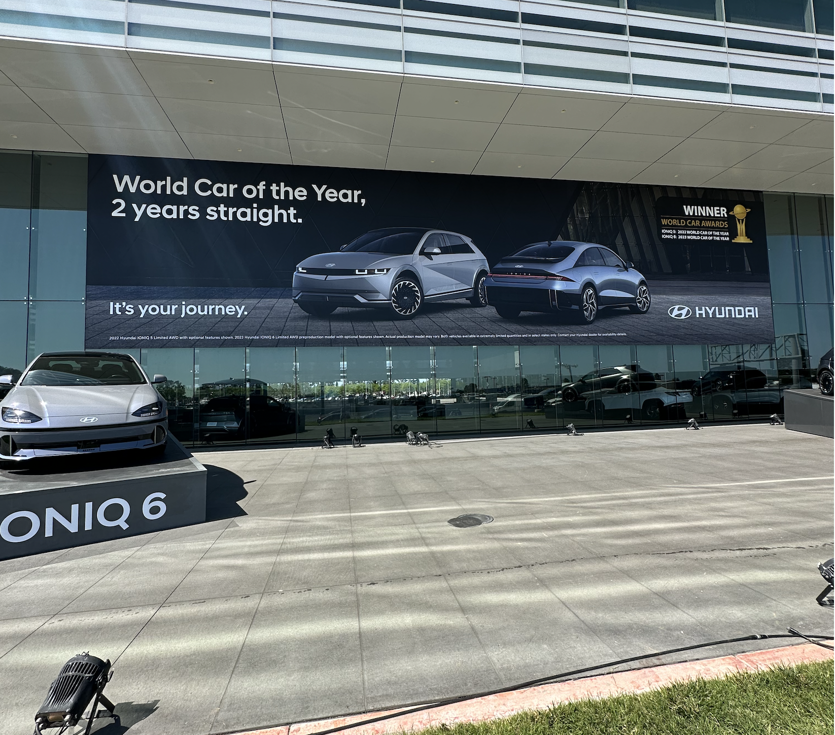 Large Hyundai billboard advertising the 2022 Hyundai Ioniq 6 as the 'World Car of the Year, 2 years straight,' featuring images of two different car models on a dealership building. The dealership displays a Hyundai Ioniq 6 in front, and the area in front of the dealership is empty.
