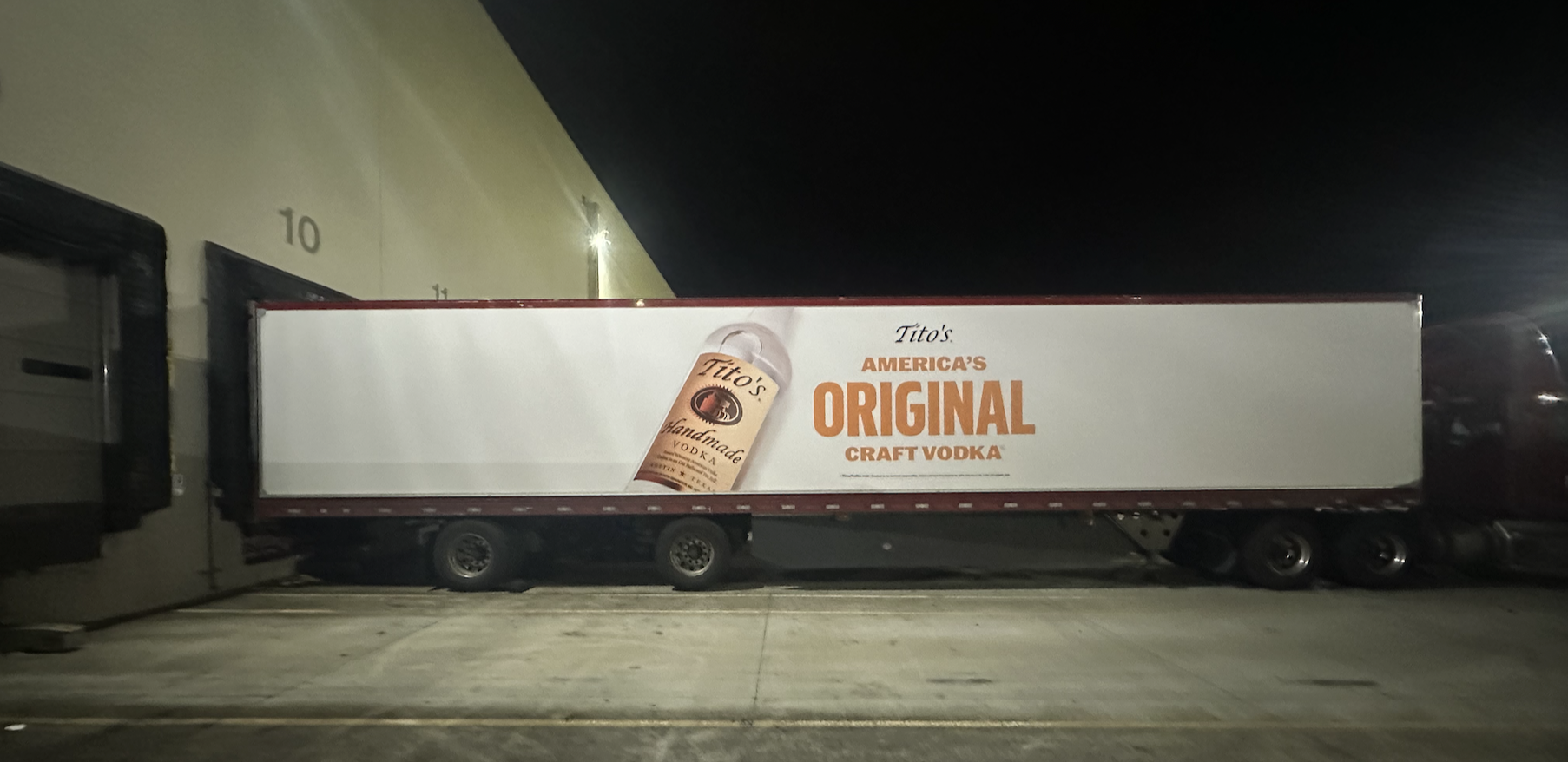 A large truck parked outside a building at night, featuring an advertisement for Tito's craft vodka, highlighting that it is America's original craft vodka.