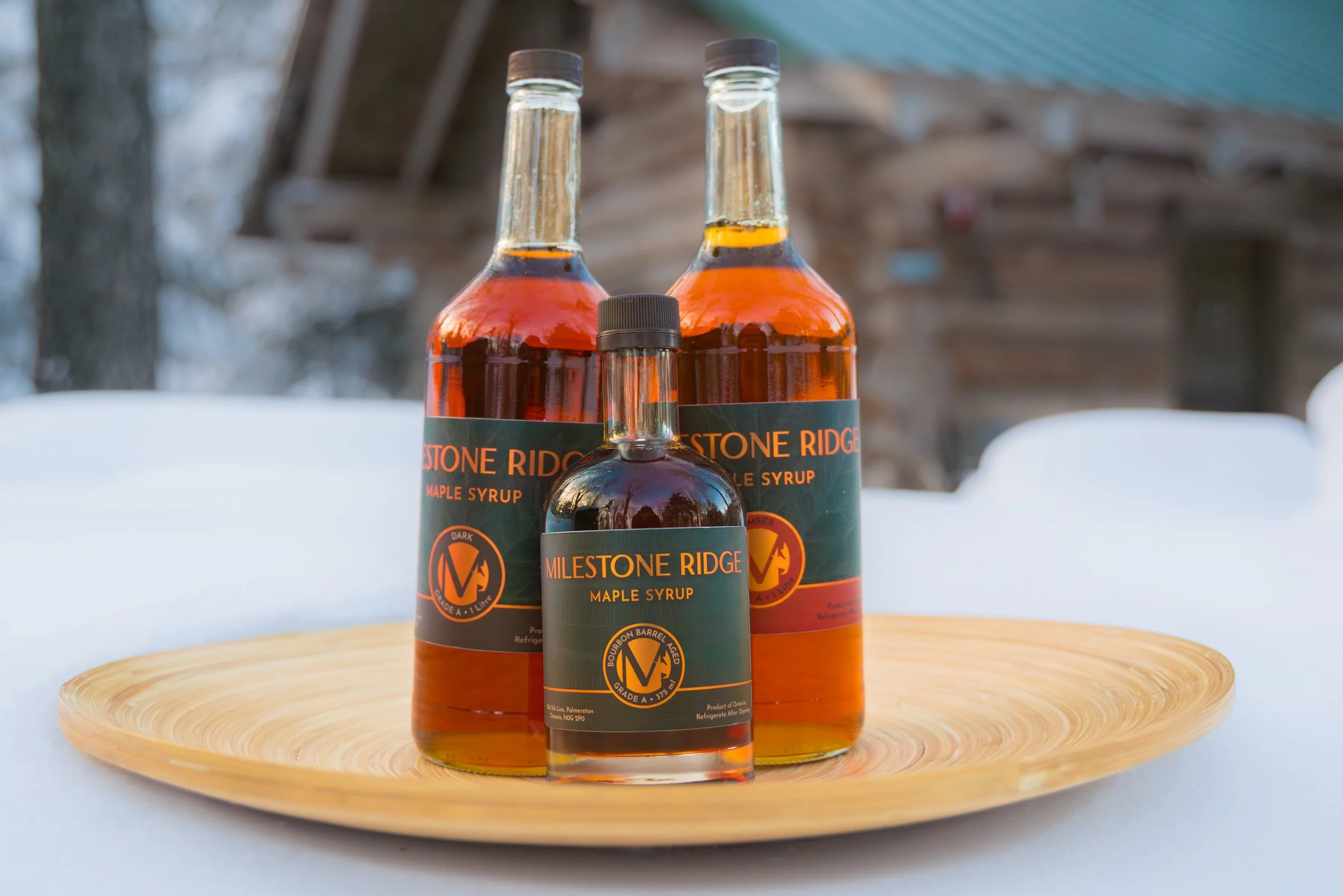 Three bottles of Milestone Ridge maple syrup on a wooden platter, with a blurred outdoor background and snow in the foreground. Wholesale maple syrup in Ontario, bulk maple syrup Canada, custom maple syrup gifts, corporate maple syrup favours.