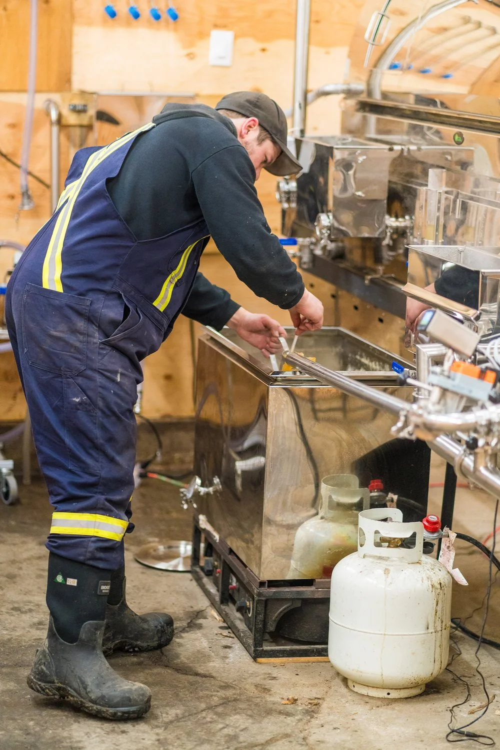 A man in work overalls and boots working with a large stainless steel tank in a workshop with tools and equipment around. Wholesale maple syrup in Ontario, bulk maple syrup Canada, custom maple syrup gifts, corporate maple syrup favours.