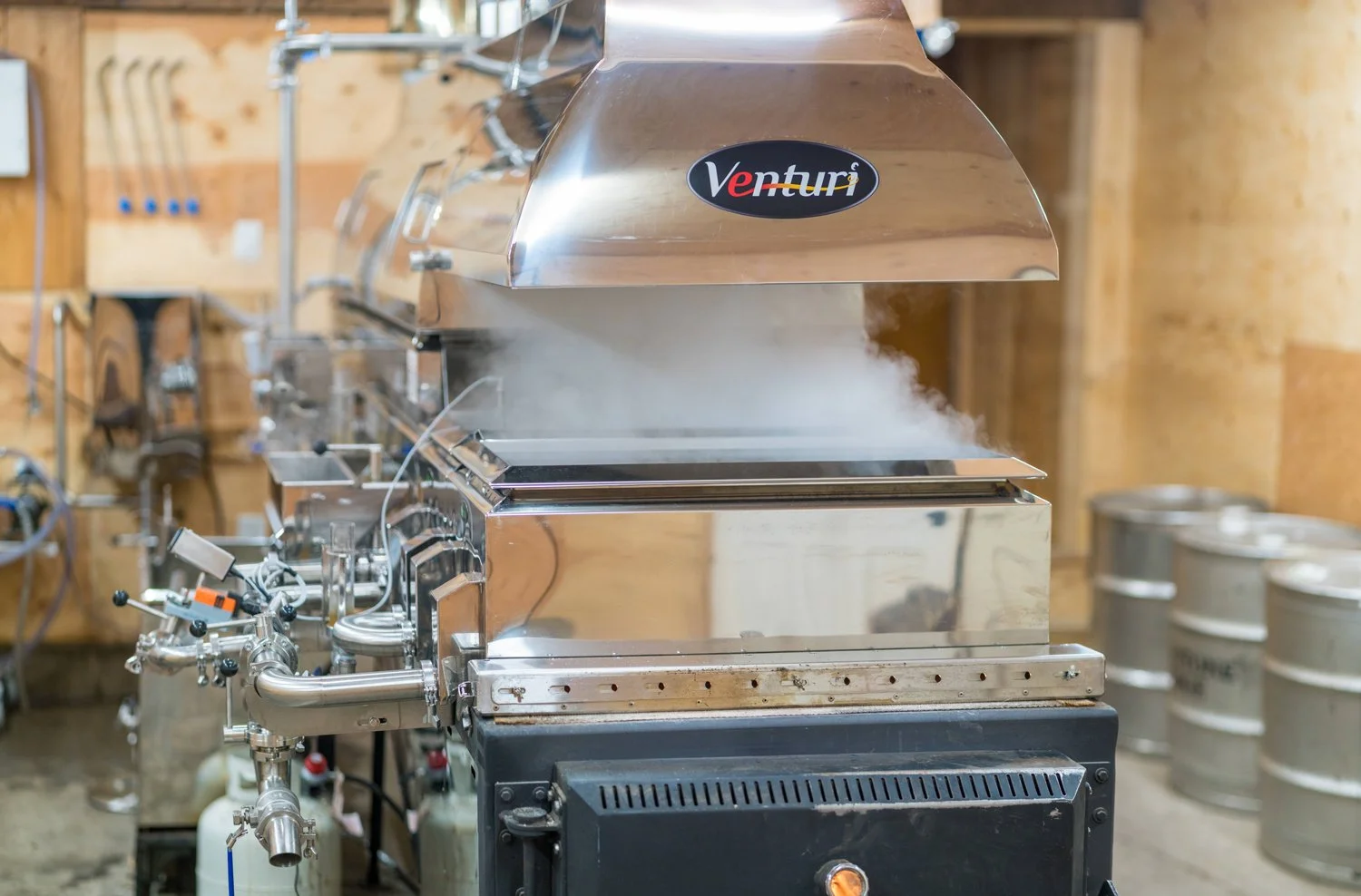 Industrial stainless steel equipment with steam or vapor coming out, labeled 'Venturi', situated in a workshop or manufacturing space with tools and barrels in the background. Wholesale maple syrup in Ontario, bulk maple syrup Canada.