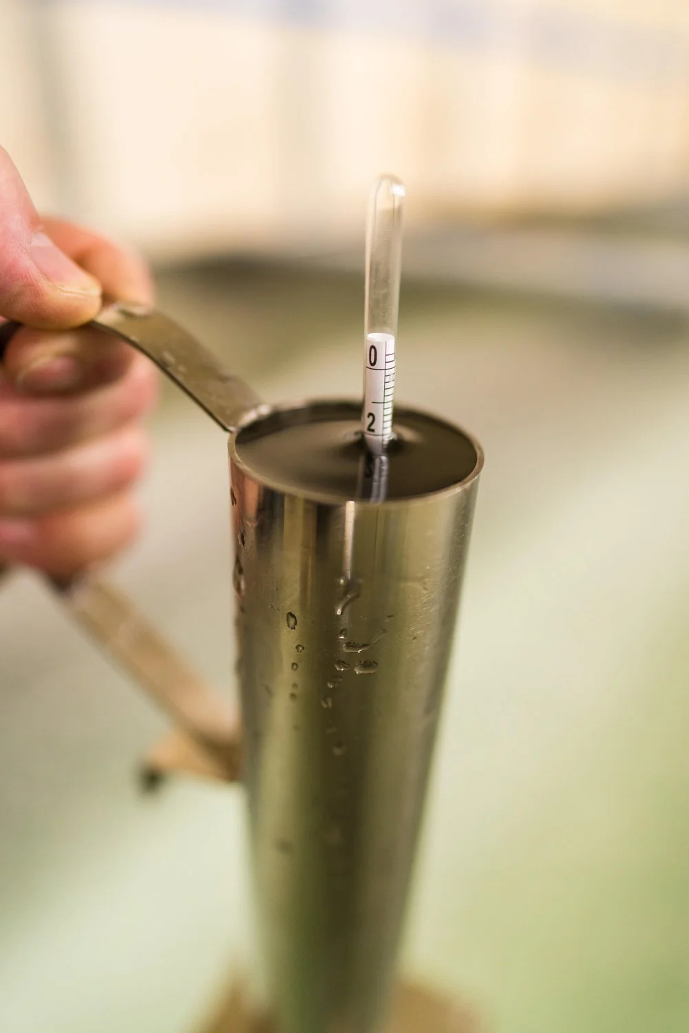 A metal container filled with maple syrup, with a thermometer partially submerged in the liquid, and a person's hand holding a tool or spatula beside the container. Milestone Ridge Maple Syrup - Pure Ontario maple syrup from Palmerston, Ontario.