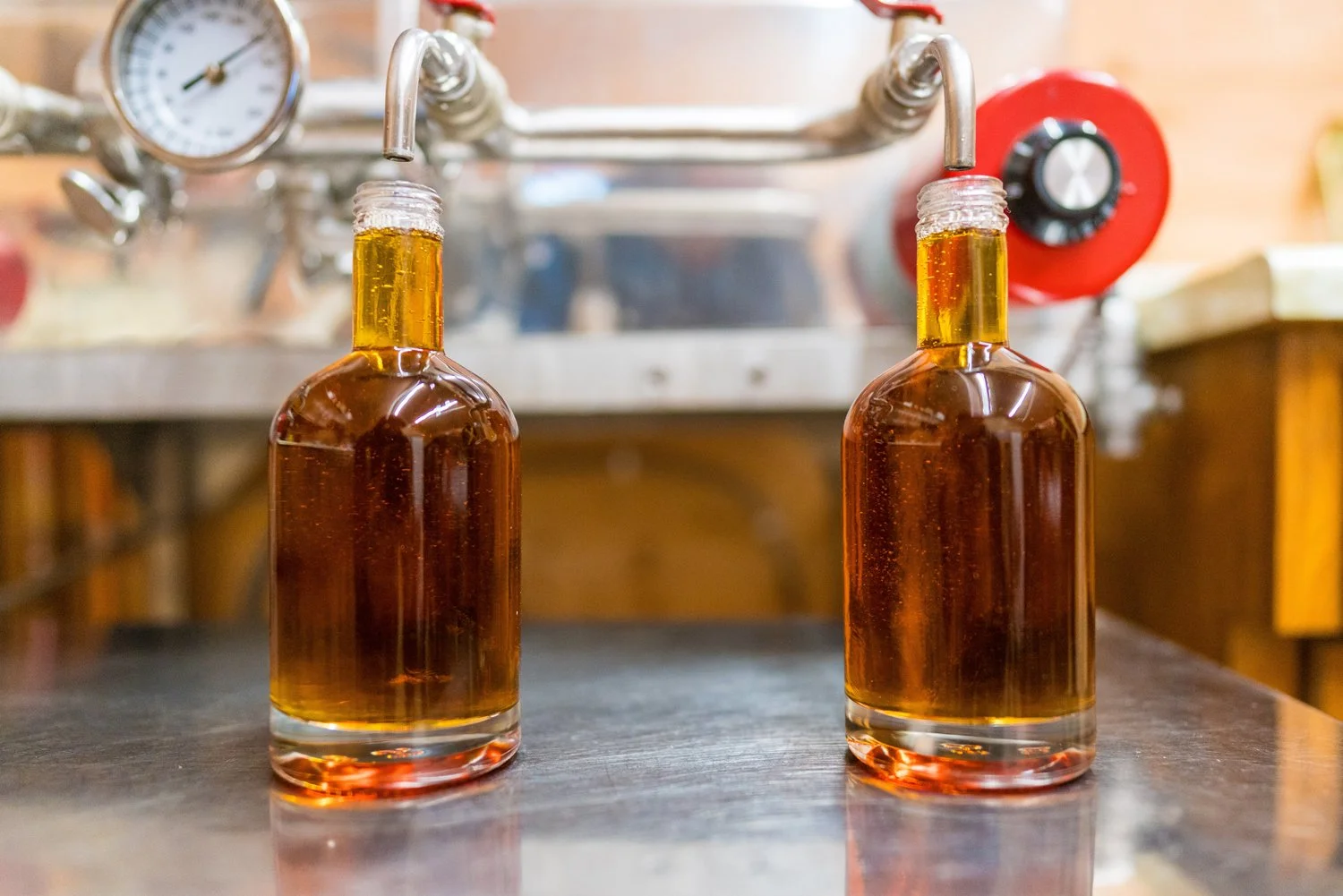 Two glass bottles filled with amber-colored liquid are connected to a brewing apparatus with metal tubes and gauges. The background shows a metal countertop and wooden cabinets.