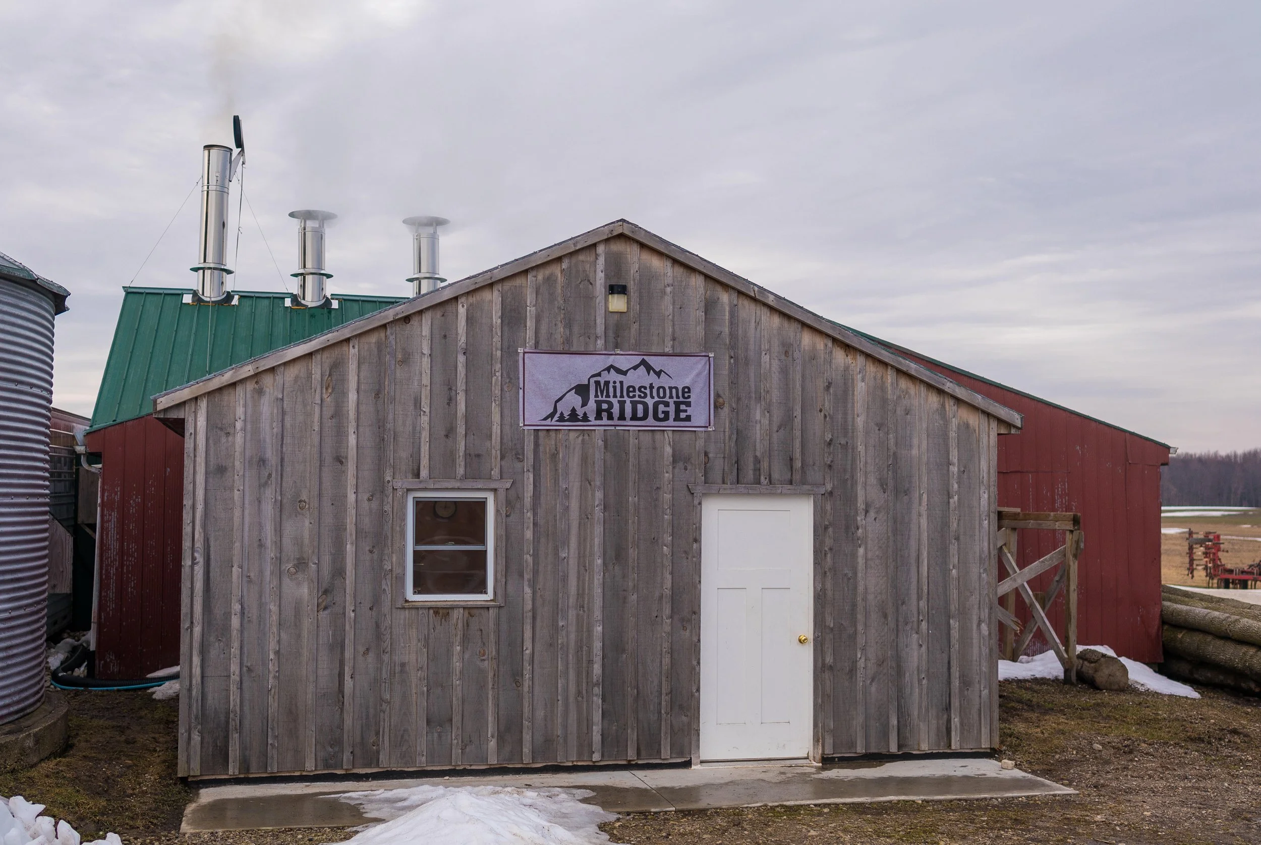 A small wooden building with a gabled roof and a sign that reads 'Milestone Ridge' on the front. The building has a white door and a window, with a green metal roof and a blue sky with some clouds overhead. Pure Ontario maple syrup from Palmerston.