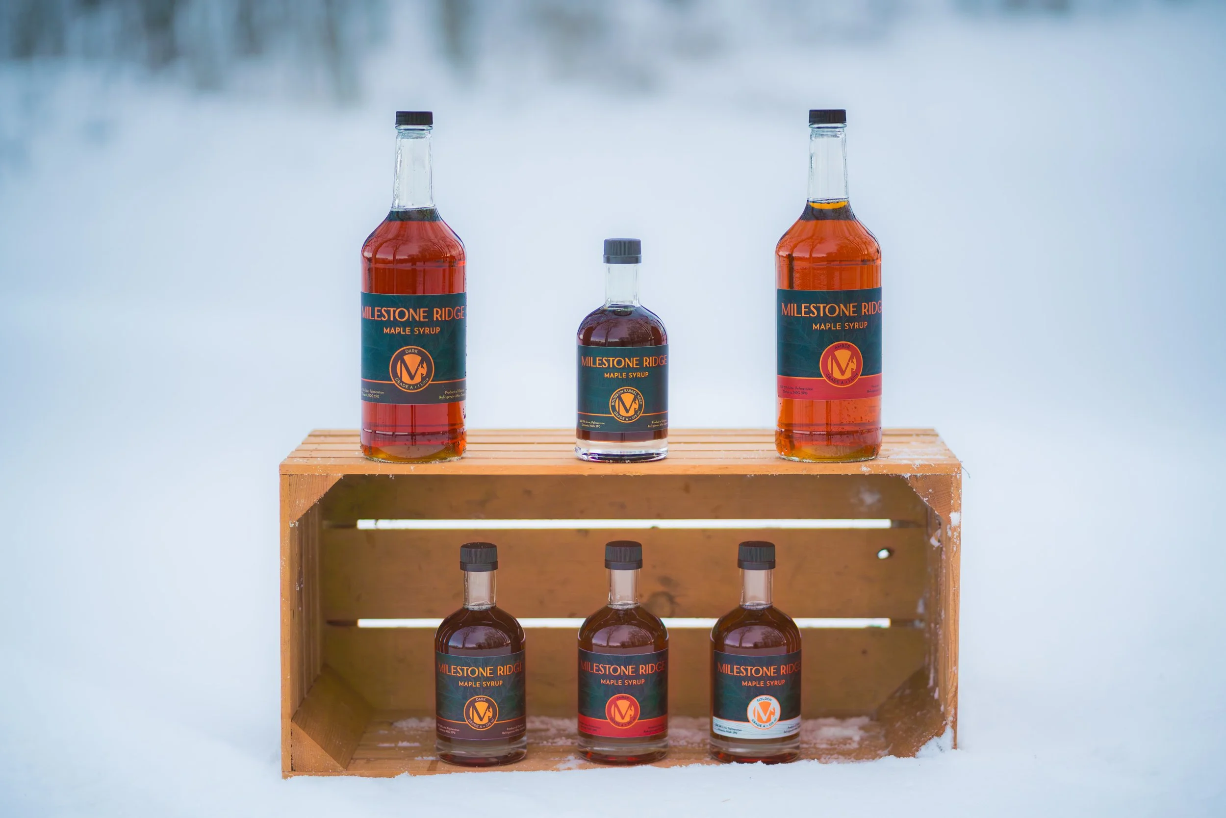 Six bottles of maple syrup positioned on a wooden display stand with a snowy background. Wholesale maple syrup in Ontario, bulk maple syrup Canada, custom maple syrup gifts, corporate maple syrup favours, maple syrup gift ideas Ontario.