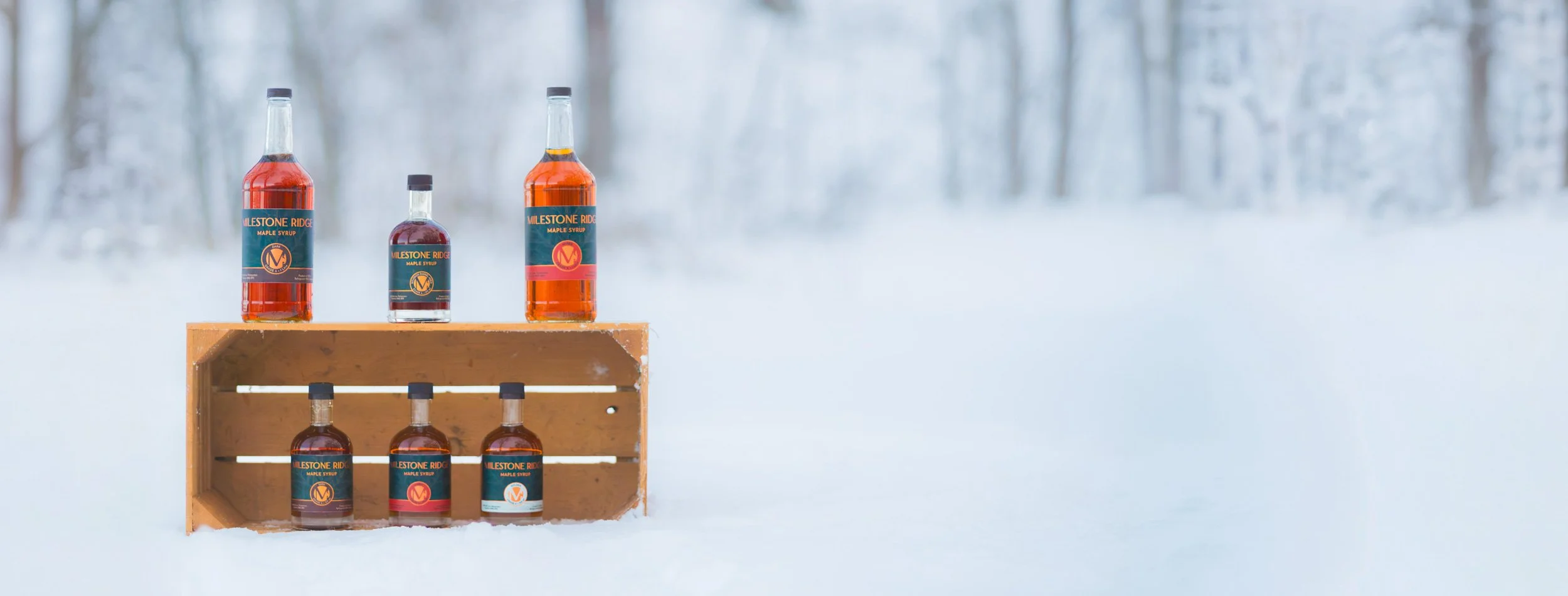 Bottles of maple syrup, labeled "Milestone Ridge Maple Syrup," arranged on a wooden crate in a snowy landscape with a blurred winter forest background. Wholesale maple syrup in Ontario, bulk maple syrup Canada, custom maple syrup gifts.