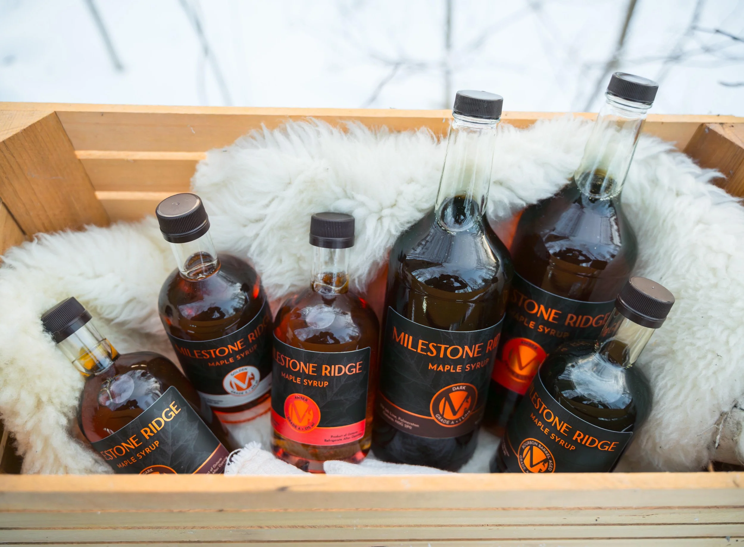 Assorted bottles of Milestone Ridge maple syrup in a wooden crate lined with white fuzzy fabric, with a snowy outdoor background. Wholesale maple syrup in Ontario, bulk maple syrup Canada, custom maple syrup gifts, corporate maple syrup favours.
