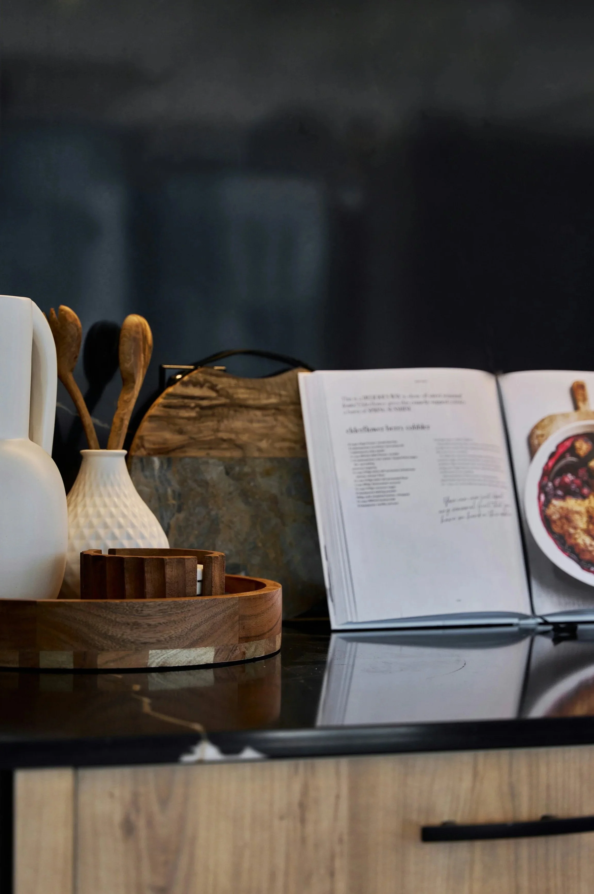 A dark table setting with white vases, wooden utensils, and an open cookbook, against a dark wall background. Grade A maple syrup available for purchase in golden maple syrup, amber maple syrup, dark maple syrup, bourbon barrel-aged maple syrup.
