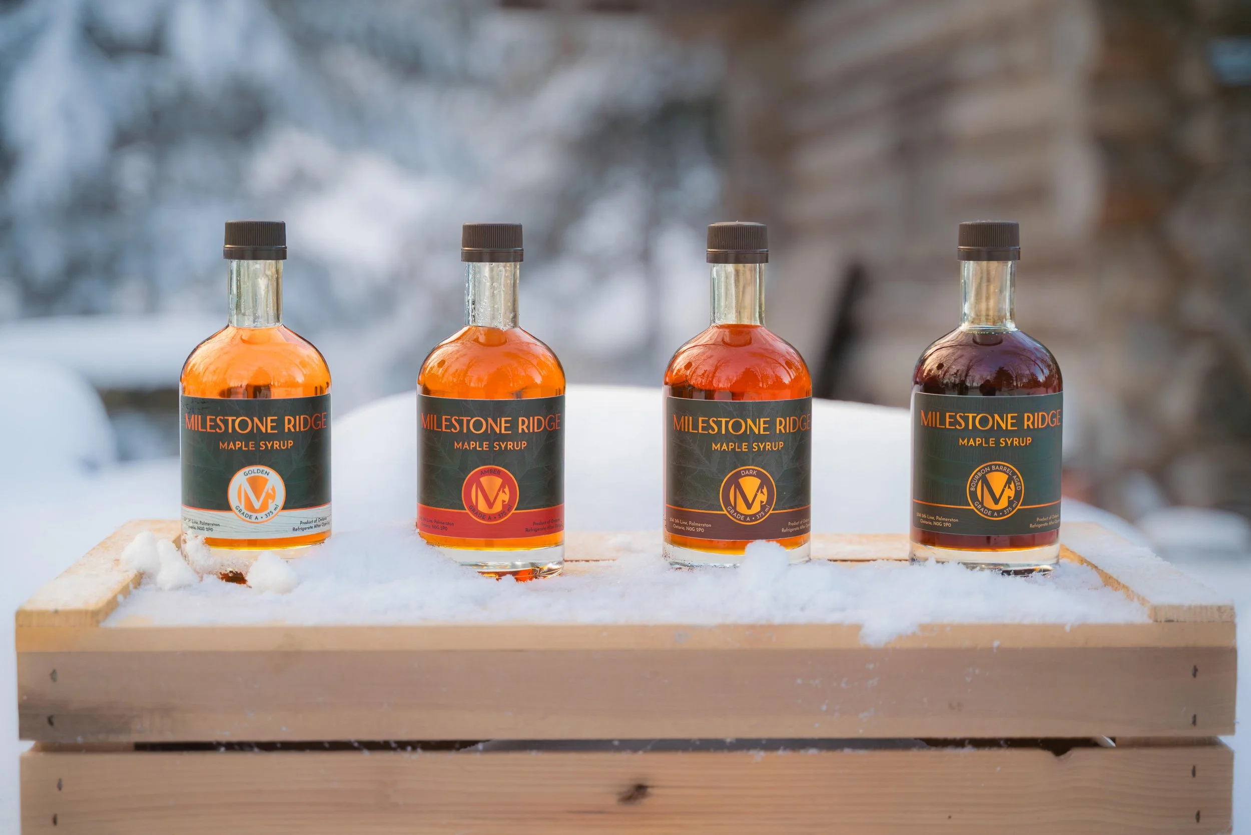 Four bottles of MileStone Ridge maple syrup in different syrup varieties placed on a wooden platform in a snowy outdoor setting. Maple syrup in Palmerston Ontario, buy maple syrup near me, maple syrup Wellington County, maple syrup Perth County.