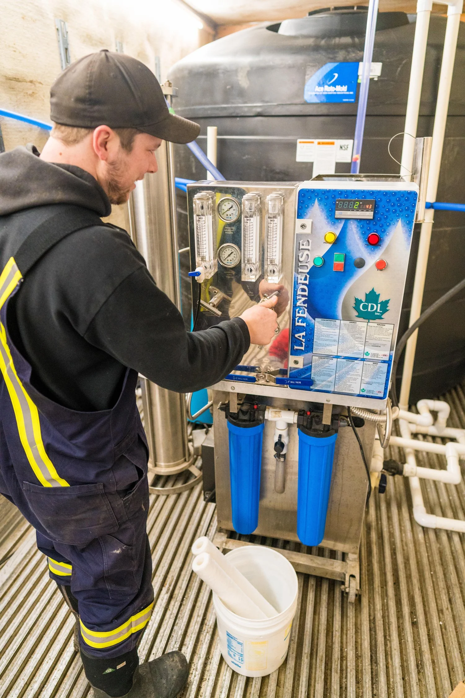 Man operating maple syrup equipment in a room. The equipment has gauges, dials, and buttons, with two large blue filters hanging from the device. The man wears work clothes and a cap, and is handling a lever or valve on the machine.